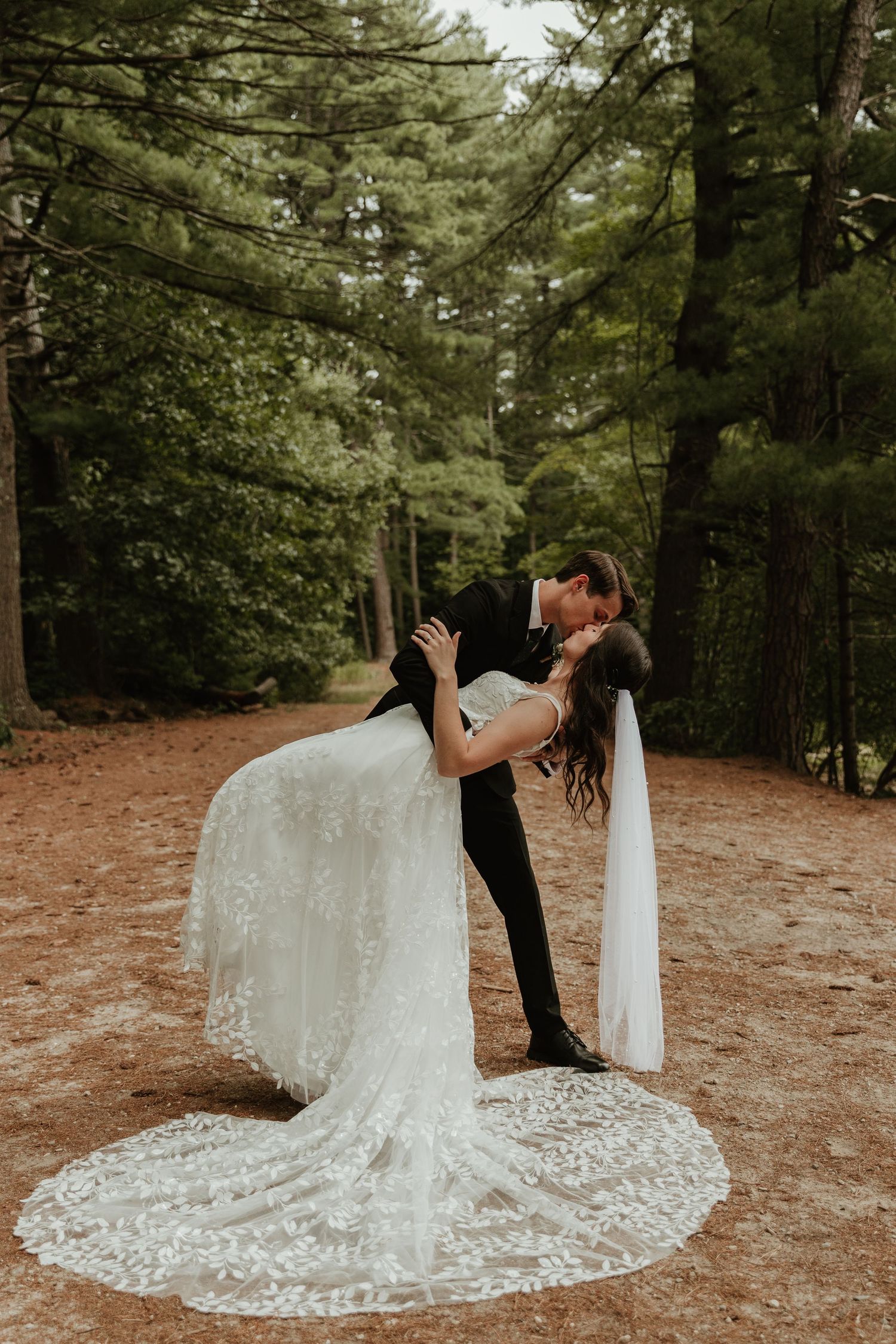 A romantic wedding couple shares a dip kiss on a forest path surrounded by tall evergreen trees.