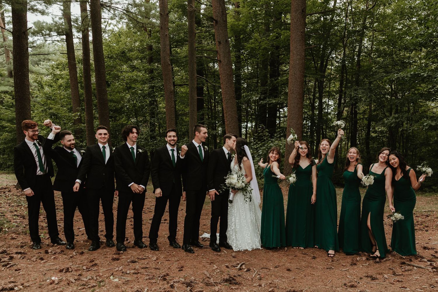 A wedding party poses playfully in a wooded setting wearing emerald green dresses and black suits.