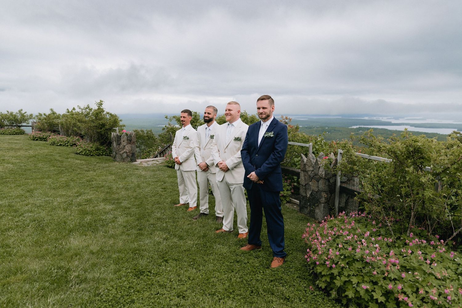 Wedding party members stand in formation on a scenic hillside overlooking a distant landscape.