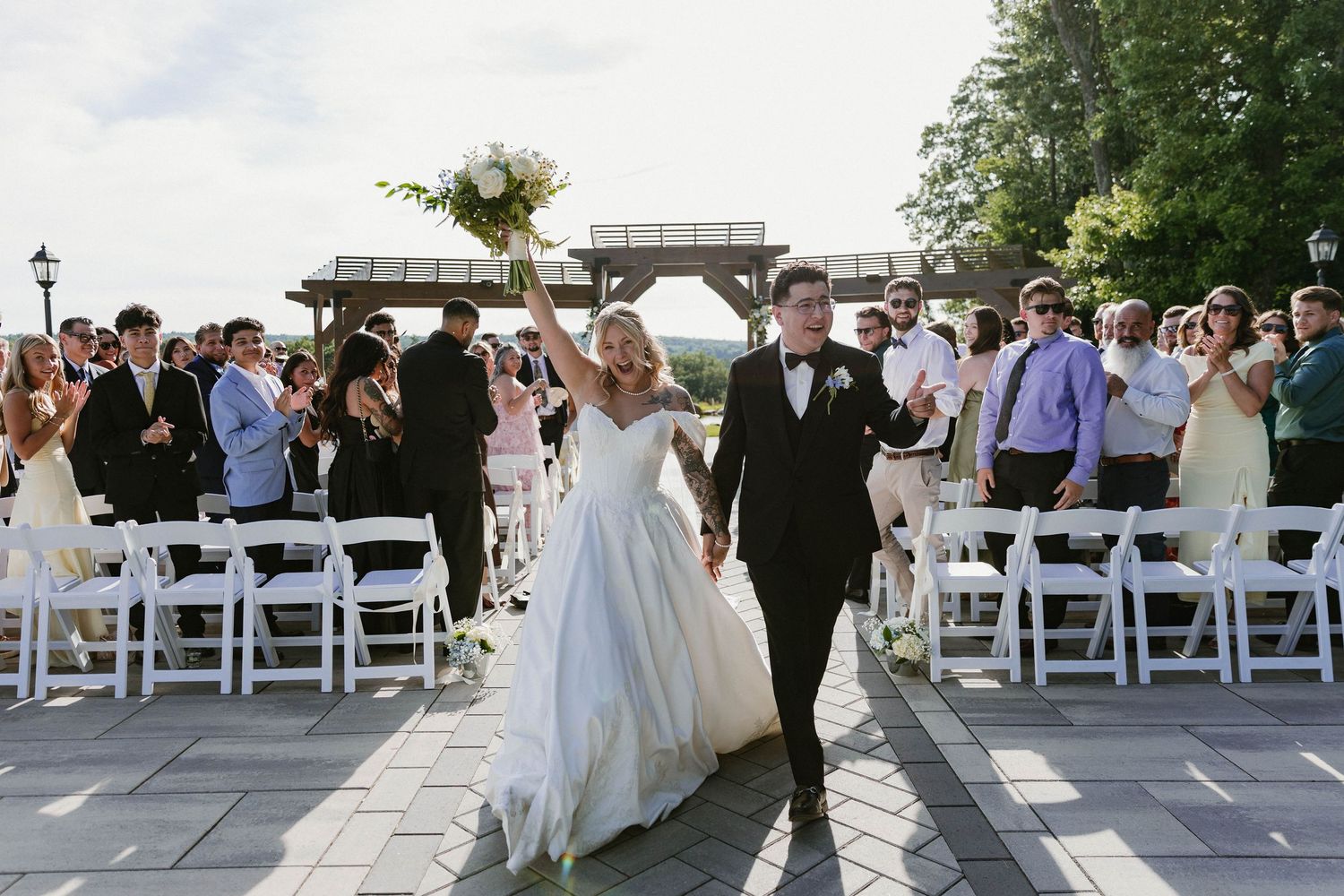 Bride and groom walk down outdoor aisle holding hands after wedding ceremony, guests clapping, white chairs, pergola backdrop.