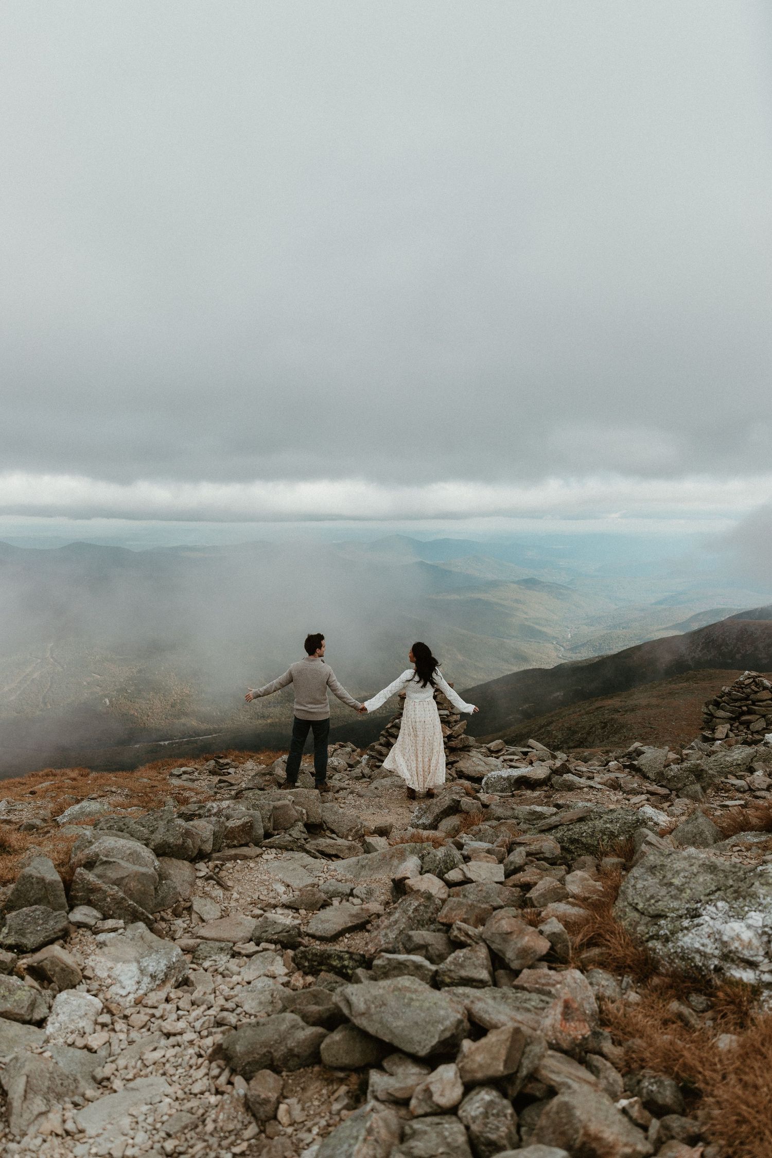 Two people stand together on a rocky mountain overlook surrounded by misty clouds.