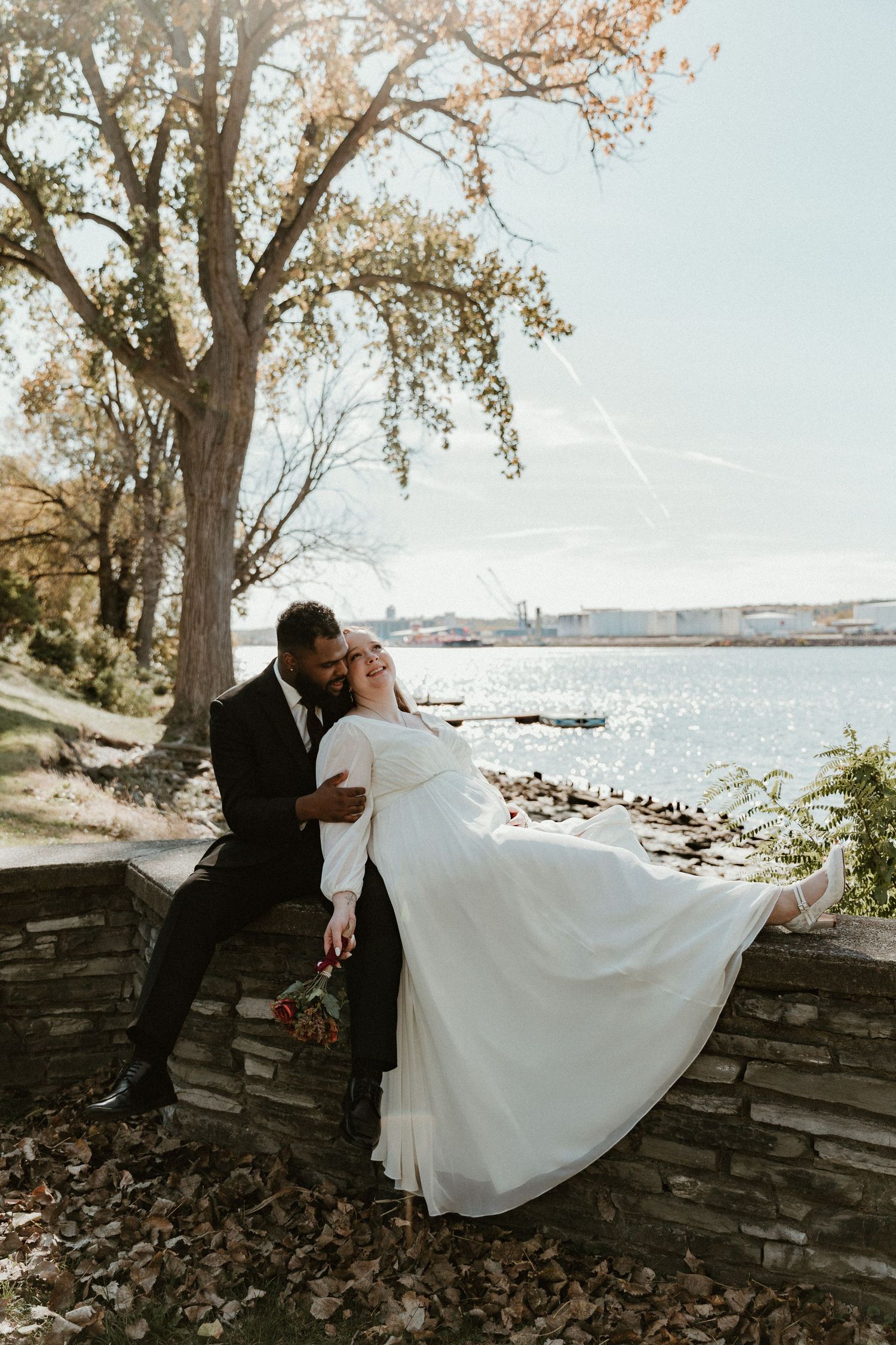 Romantic wedding photo of couple by waterfront stone wall in autumn setting.