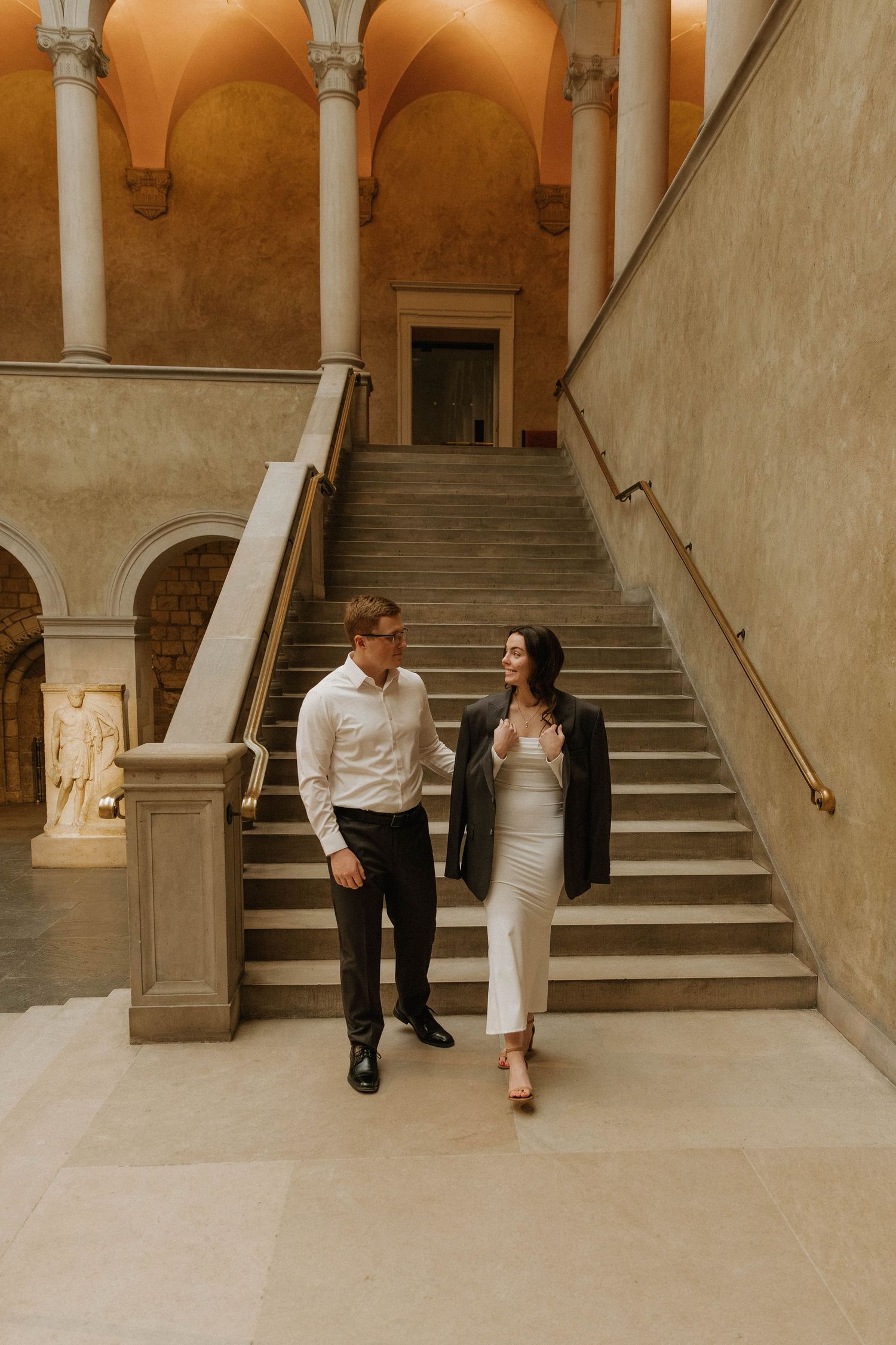 A couple in formal attire walks through an elegant archway with marble columns and stairs in a historic building.