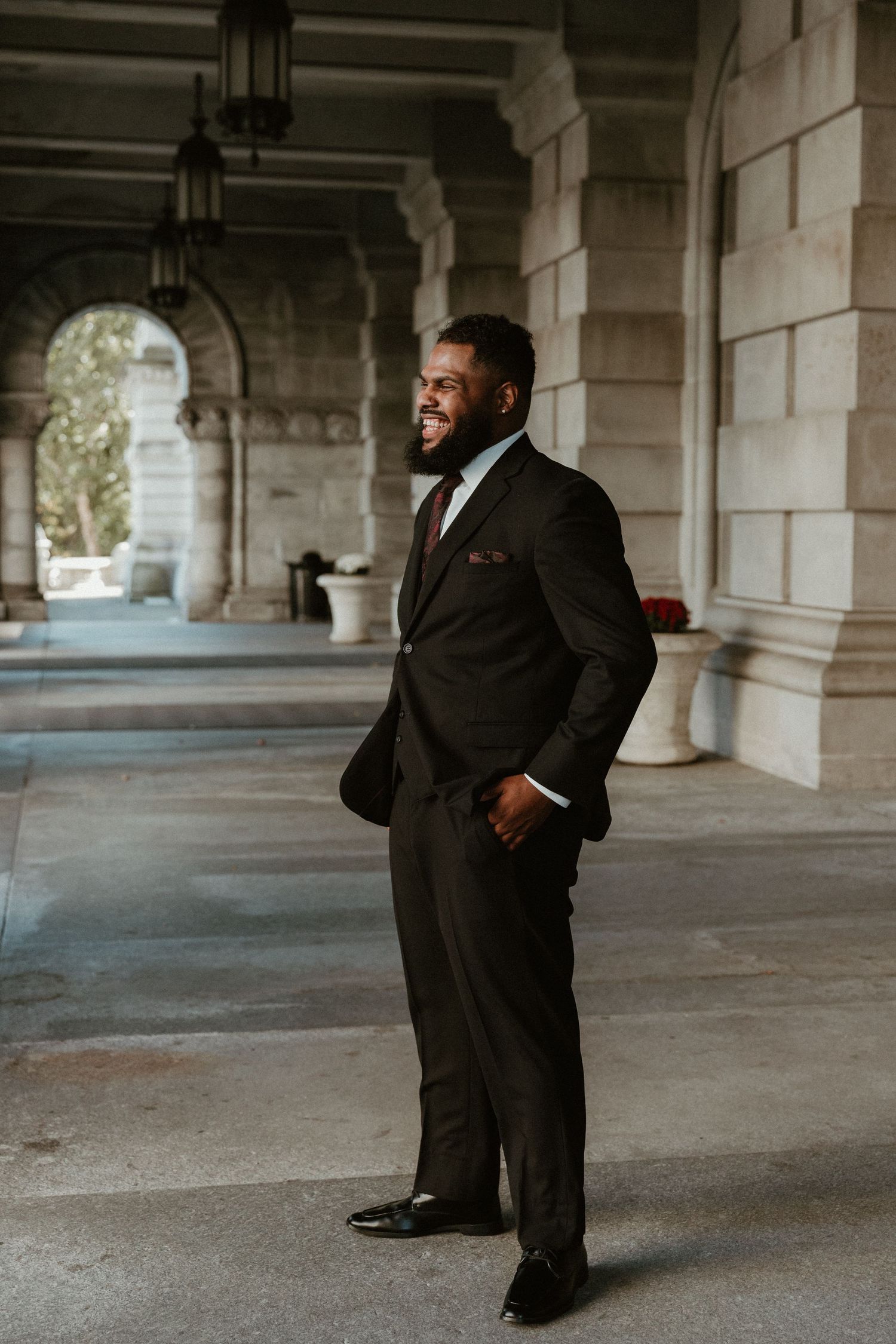 Groom in black suit smiles while standing in architectural colonnade.