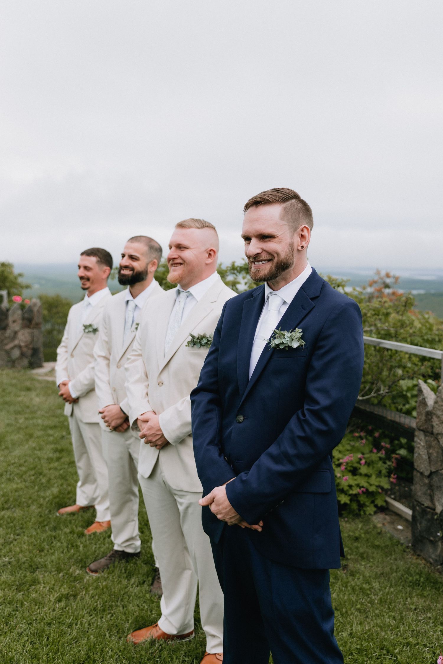 Groomsmen in white suits and navy blue stand together on a grassy hilltop during an outdoor wedding ceremony.