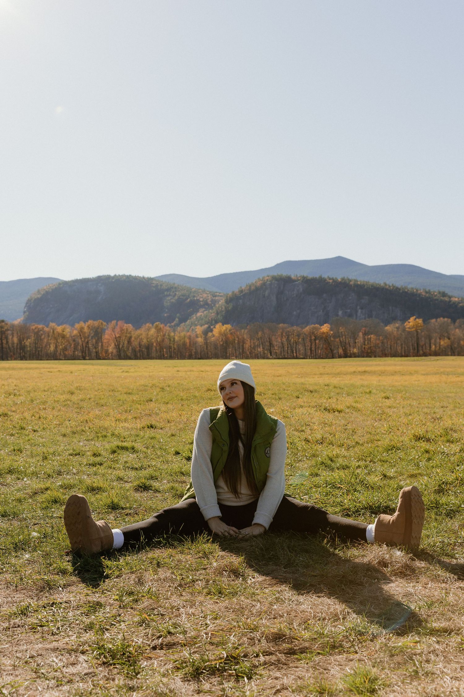 A person in a brown vest stretches on a grassy field with mountains and autumn trees in the background.