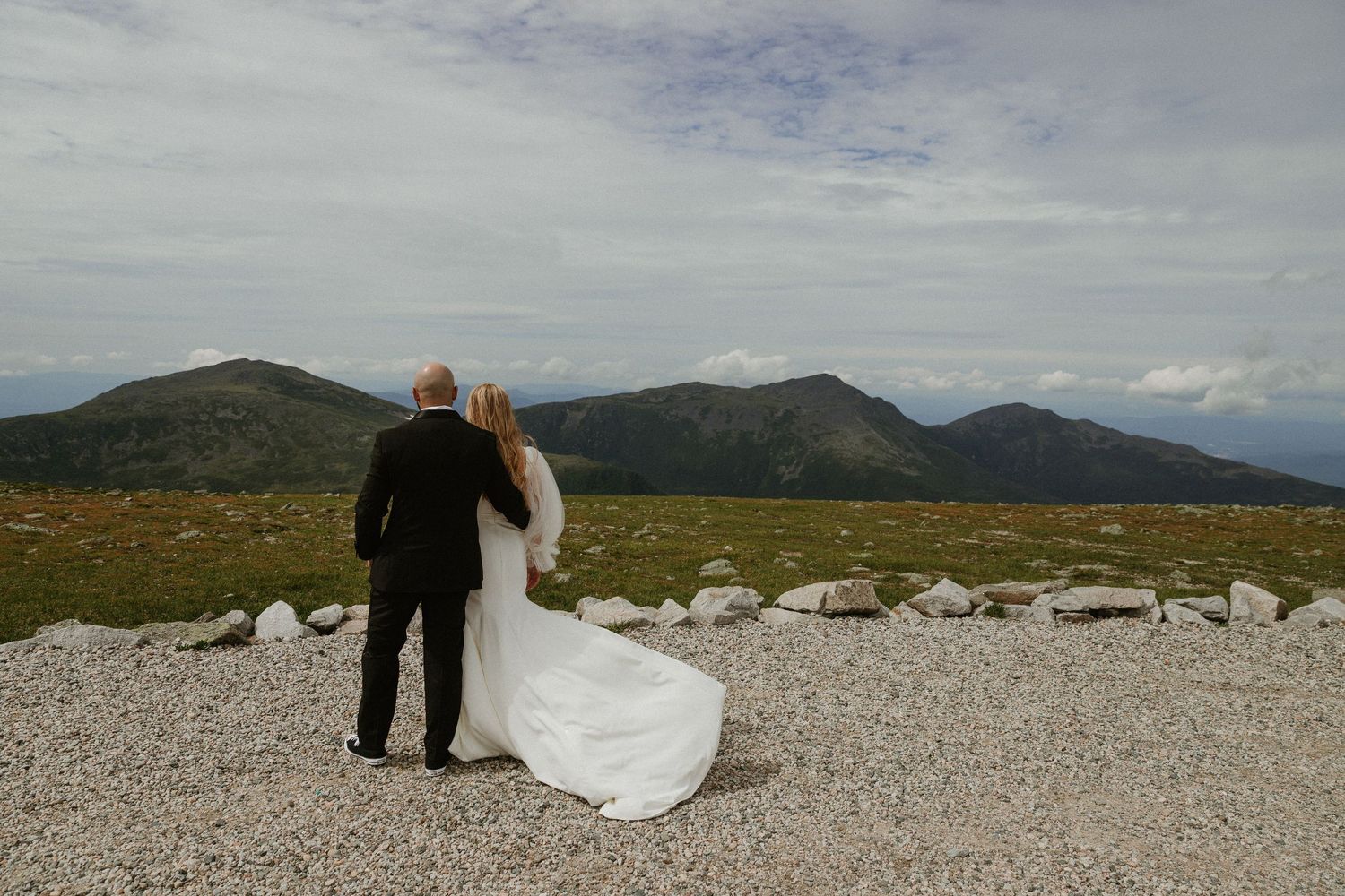 Couple in wedding attire standing on rocky mountain terrain with scenic views.