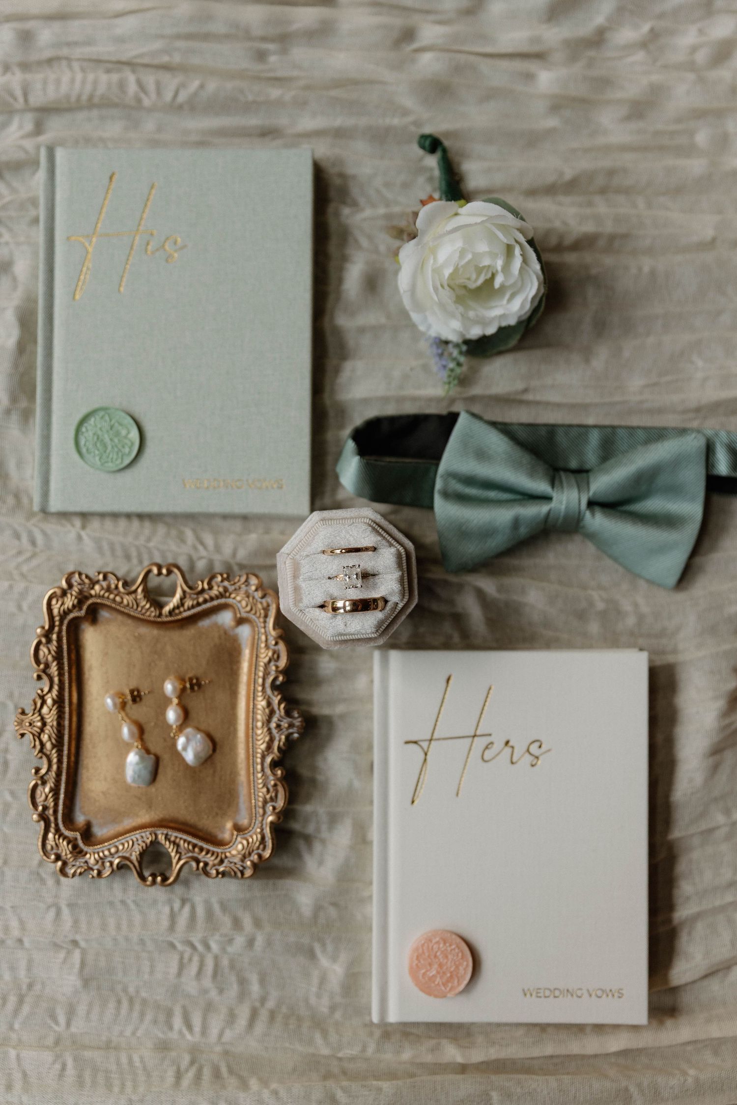 Elegant wedding flat lay with sage green bow tie, vow books, pearl earrings, rings, and floral boutonniere on linen.