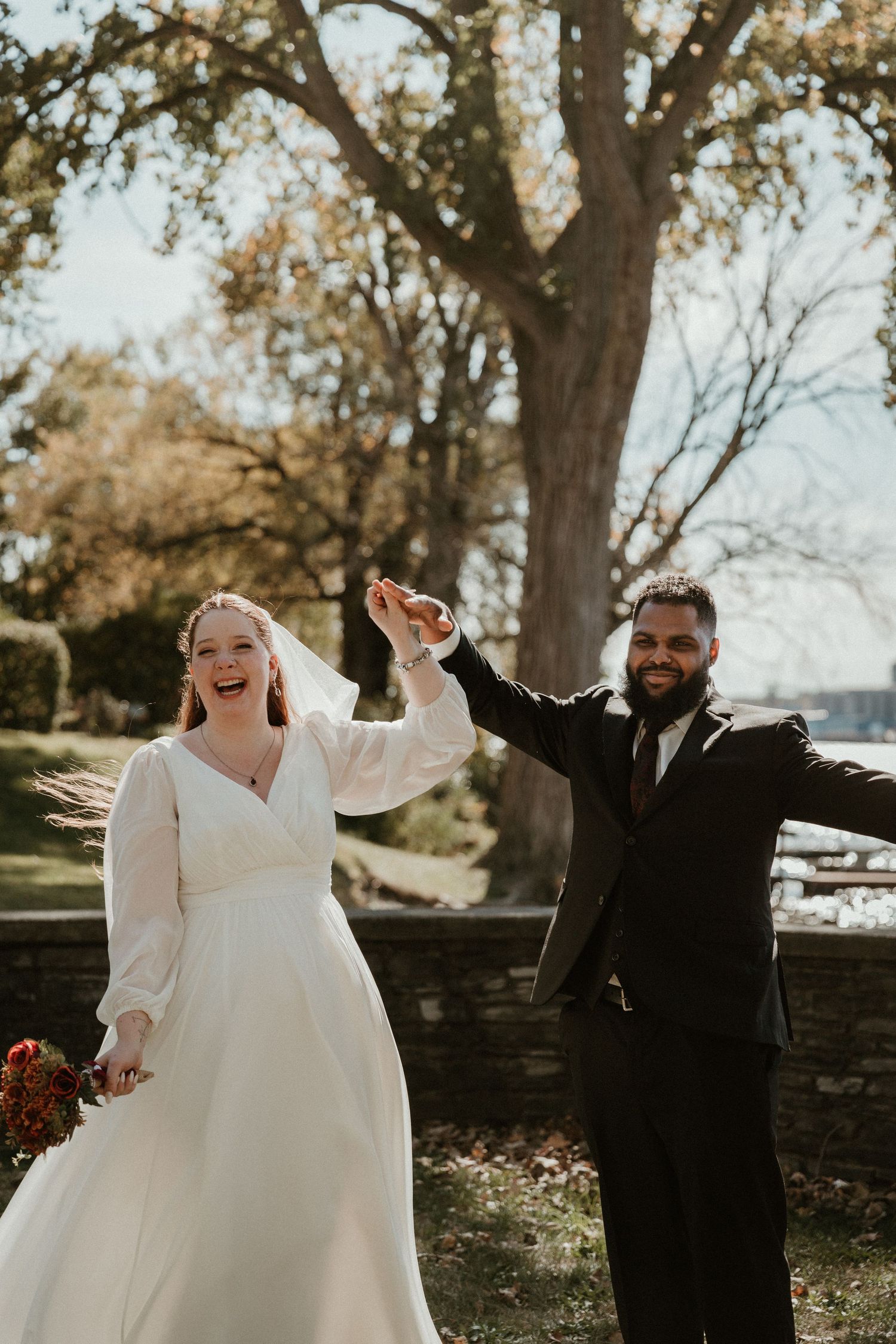 A couple celebrates their wedding day with raised hands and laughter under a majestic tree.