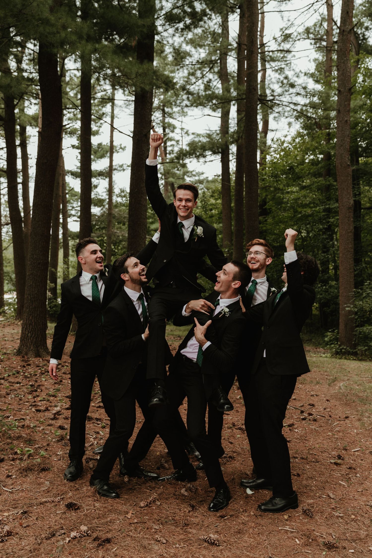 Groomsmen in black suits pose playfully in a wooded forest setting for wedding photos.
