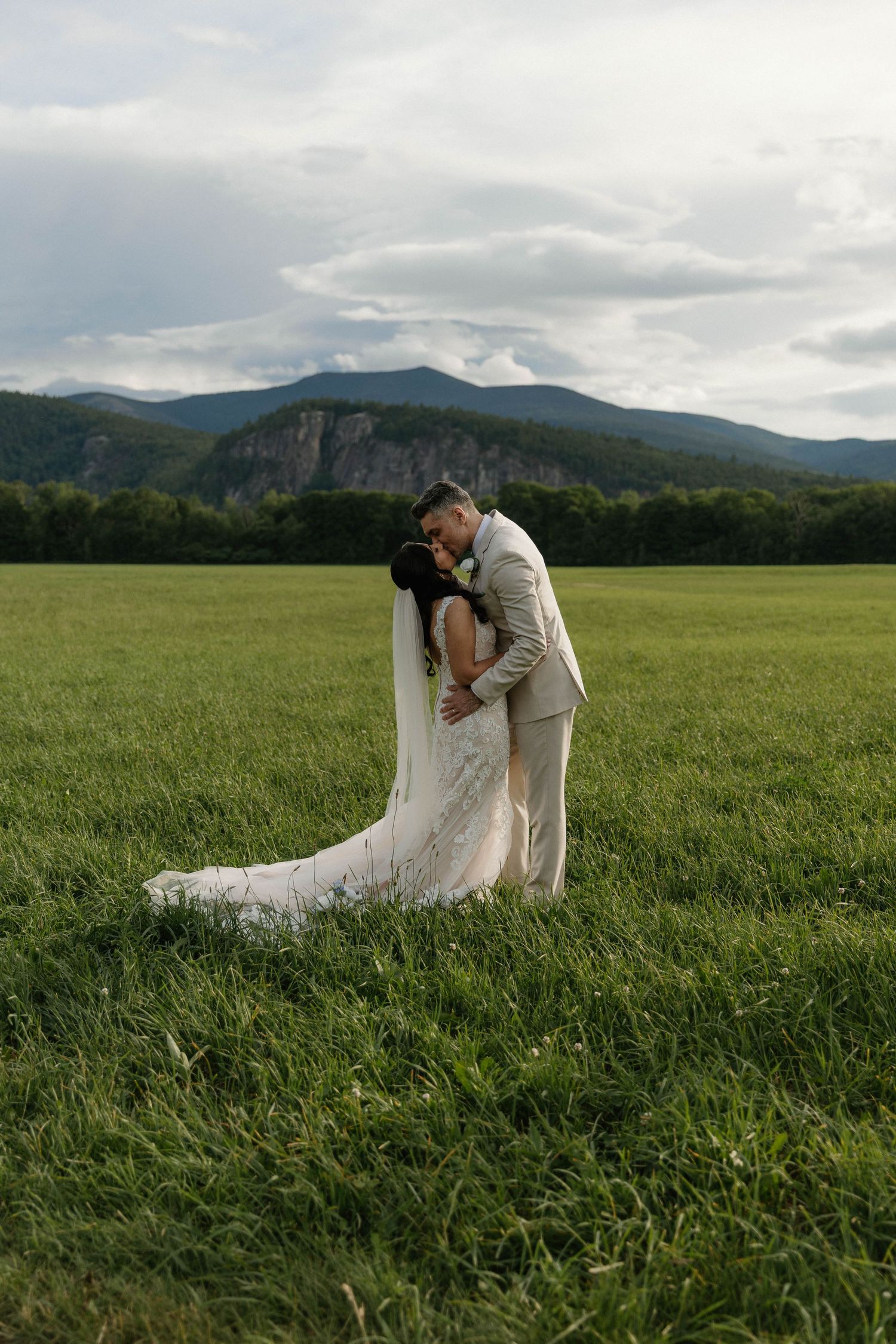 Bride and groom embrace in a lush green field with mountain backdrop on their wedding day.