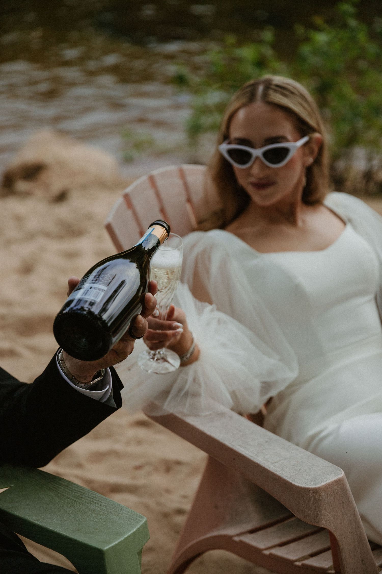 A couple pours and clinks champagne glasses in celebration at a beach setting.