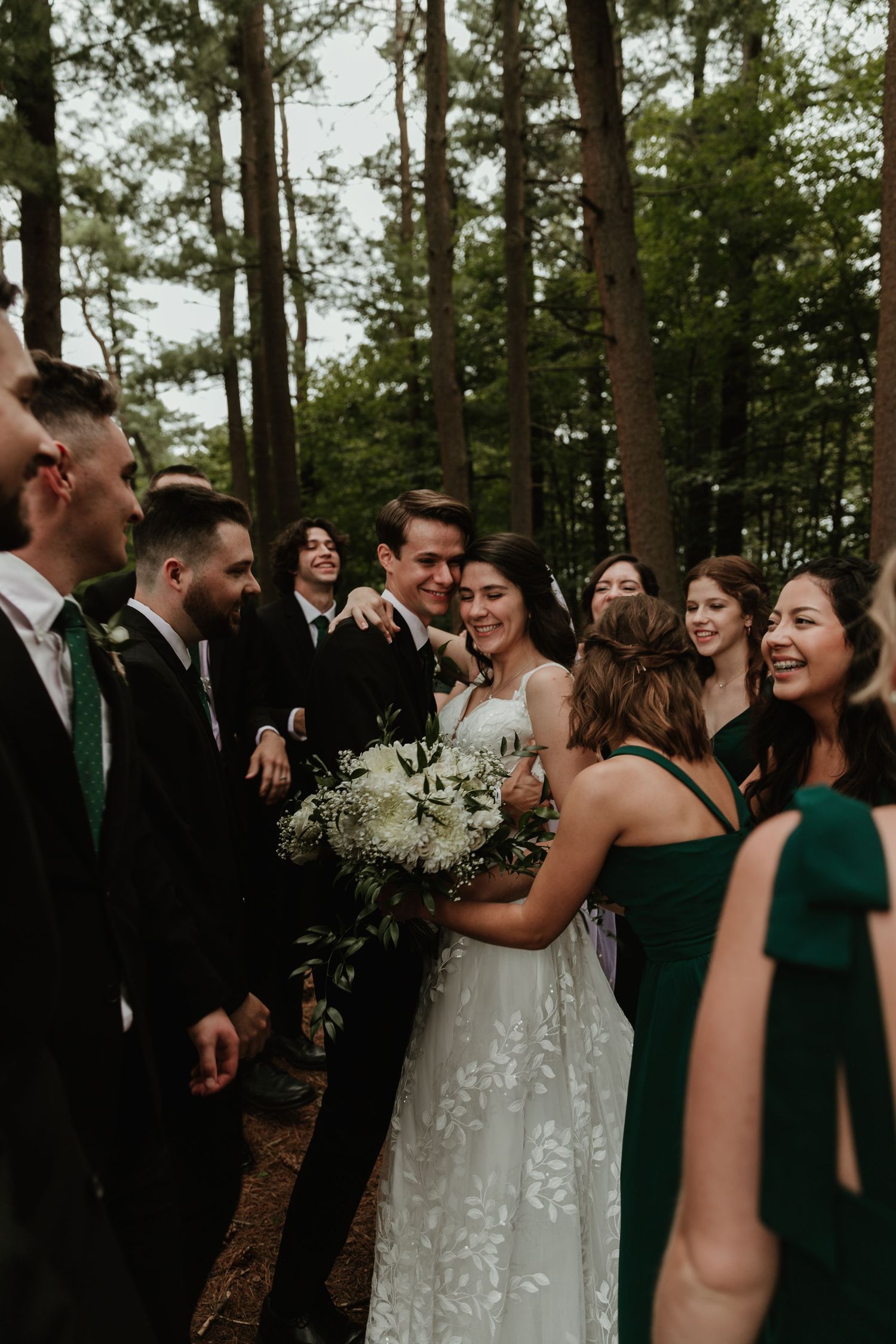 Wedding party in forest setting wearing emerald green dresses and suits share joyful moments among pine trees.