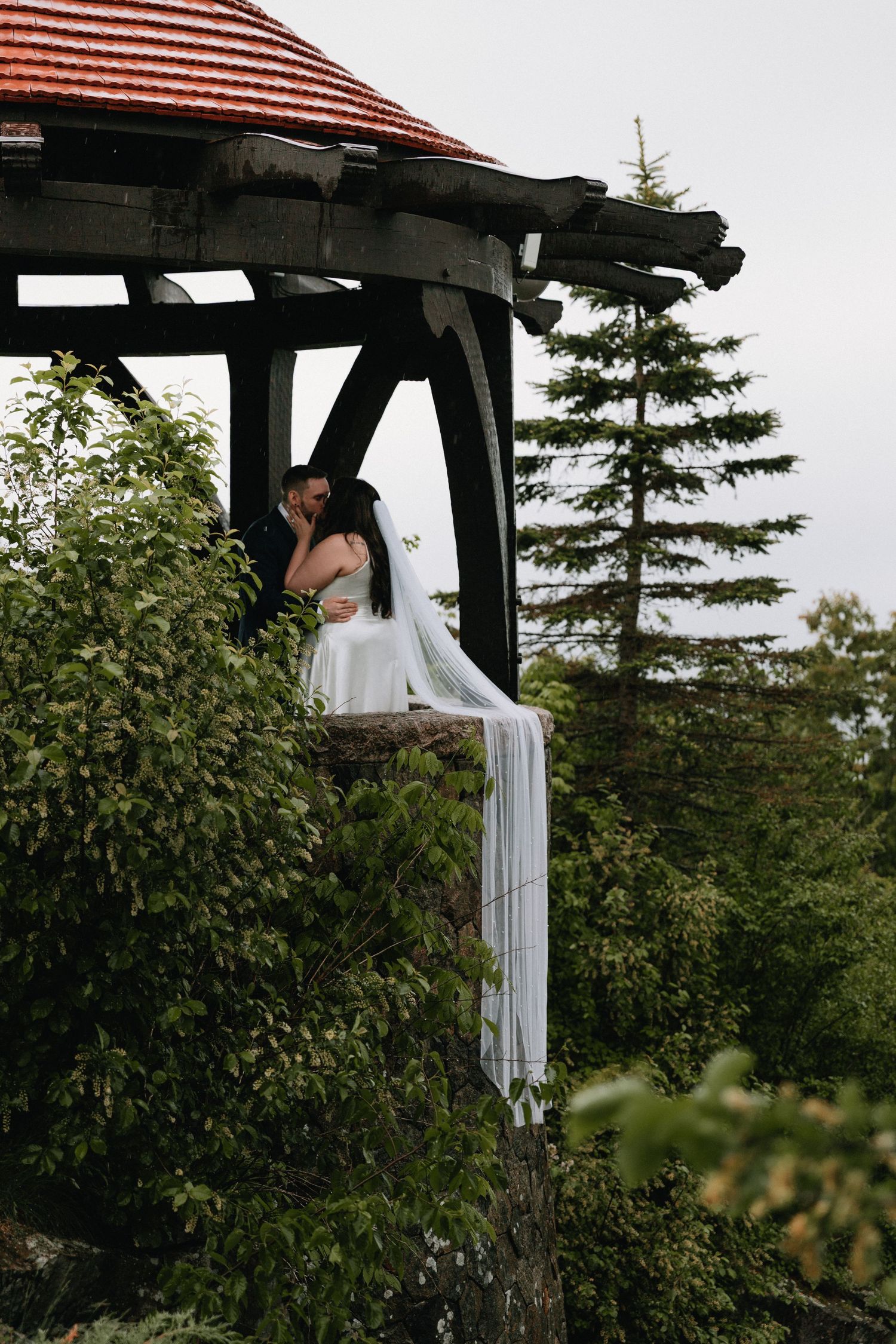 White fabric draping elegantly from a wooden pergola structure against a backdrop of evergreen trees.
