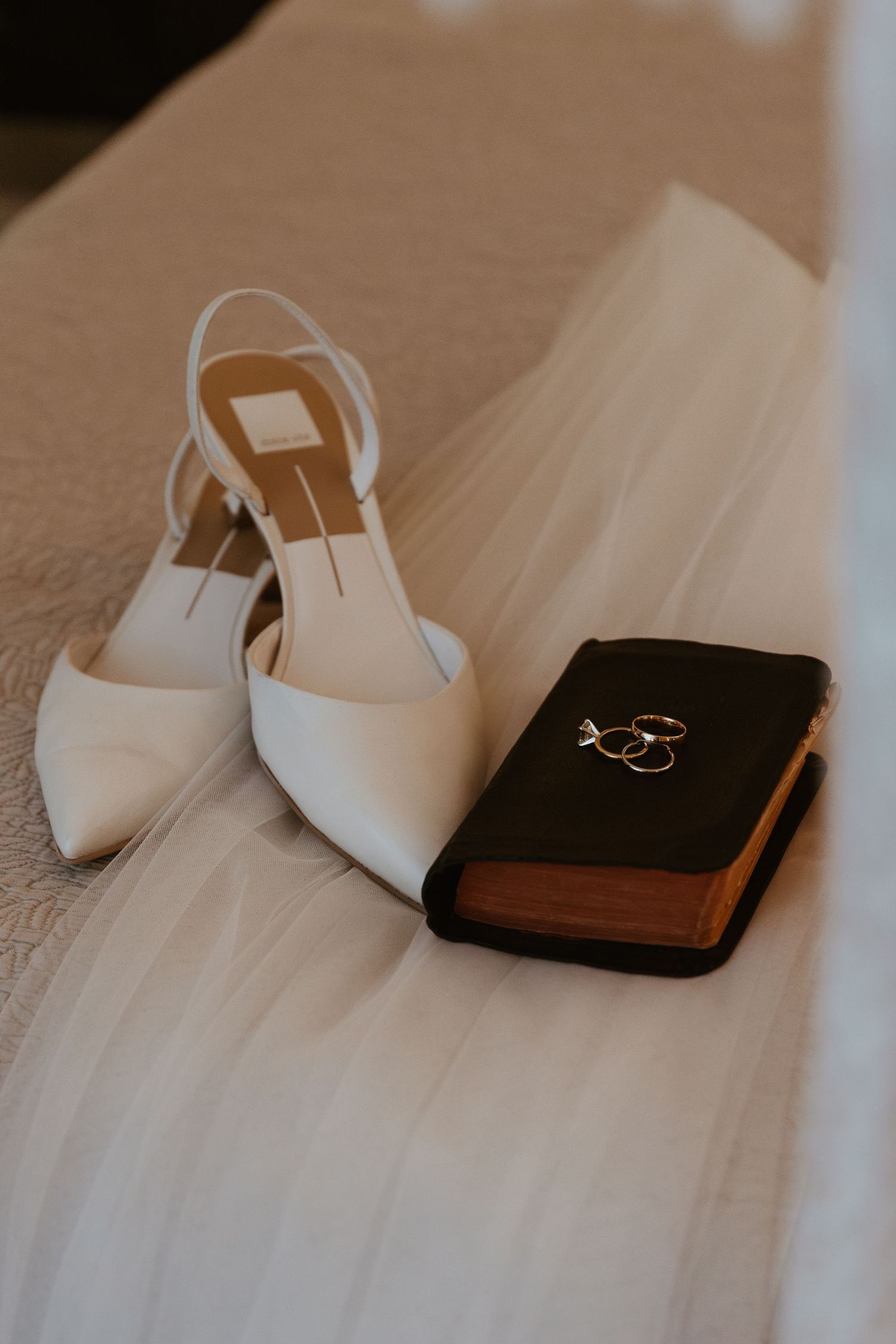 White bridal heels and a brown leather wallet displayed on a beige surface.