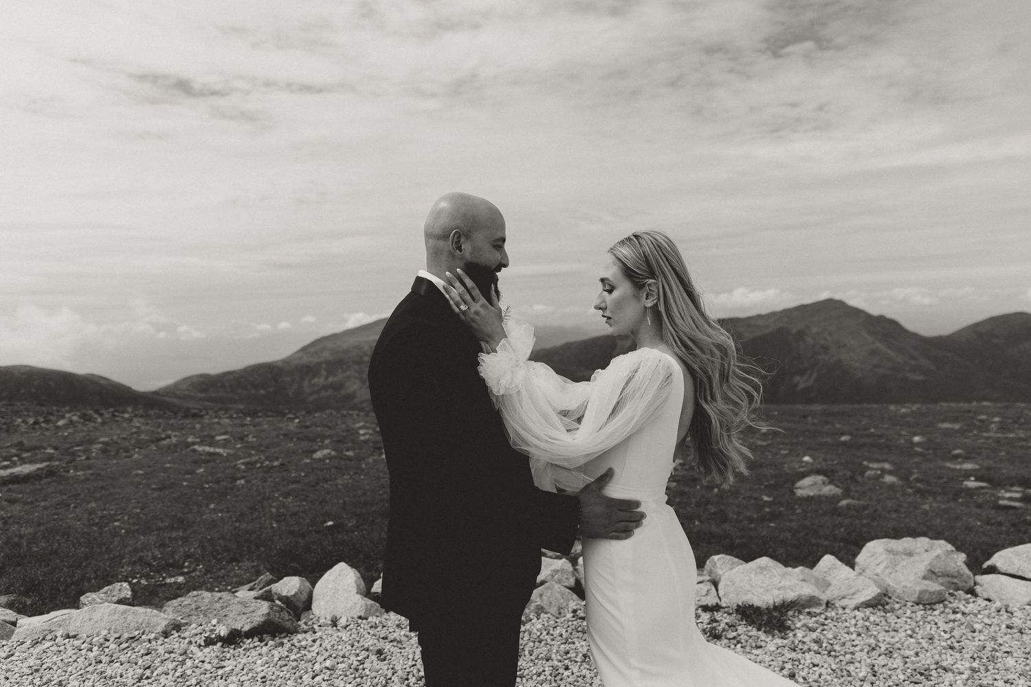 A wedding couple shares an intimate moment atop a scenic mountain vista with dramatic cloudy skies.