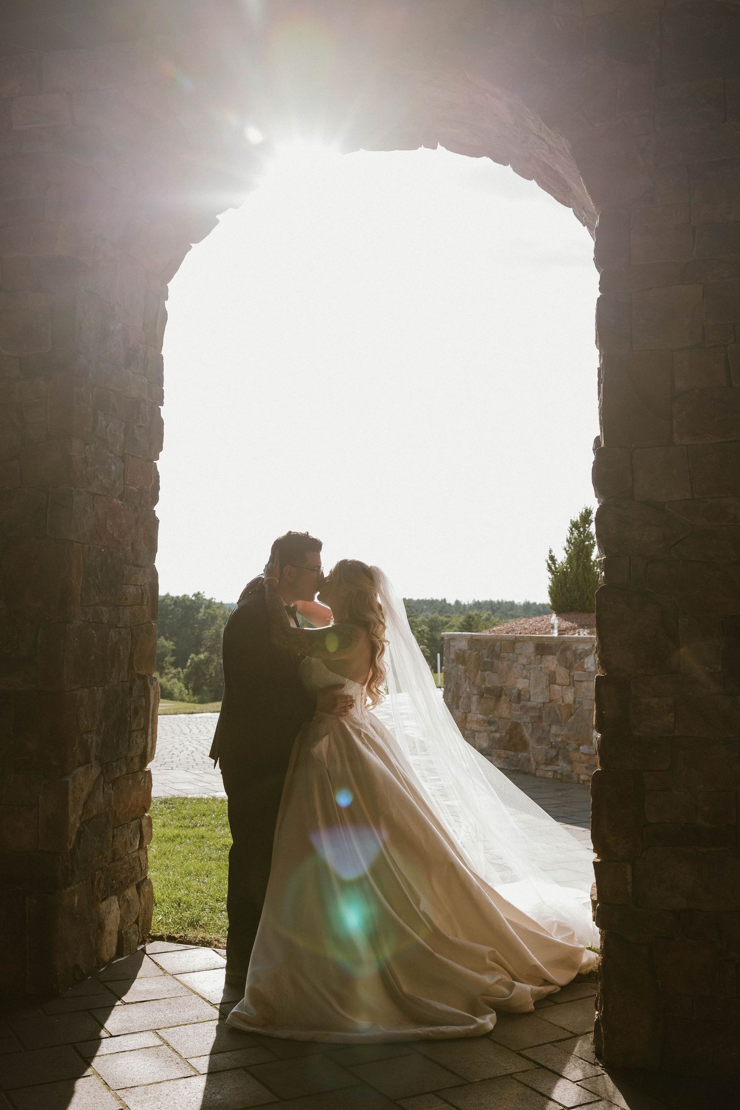 Bride and groom silhouetted in a stone archway with sun flare, wedding veil flowing, romantic outdoor ceremony.
