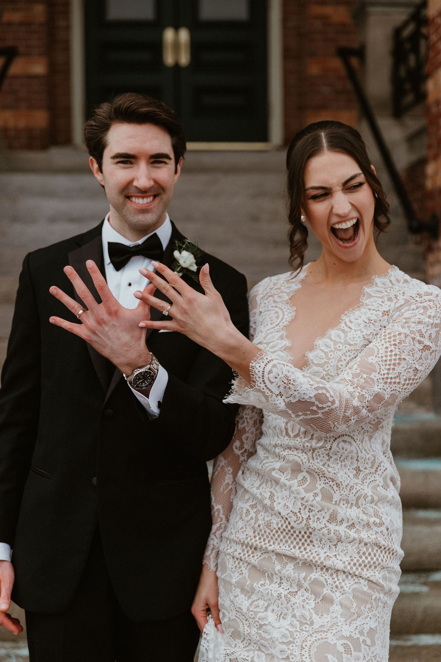 Newlyweds showing off their rings with joyful expressions outside a brick building.