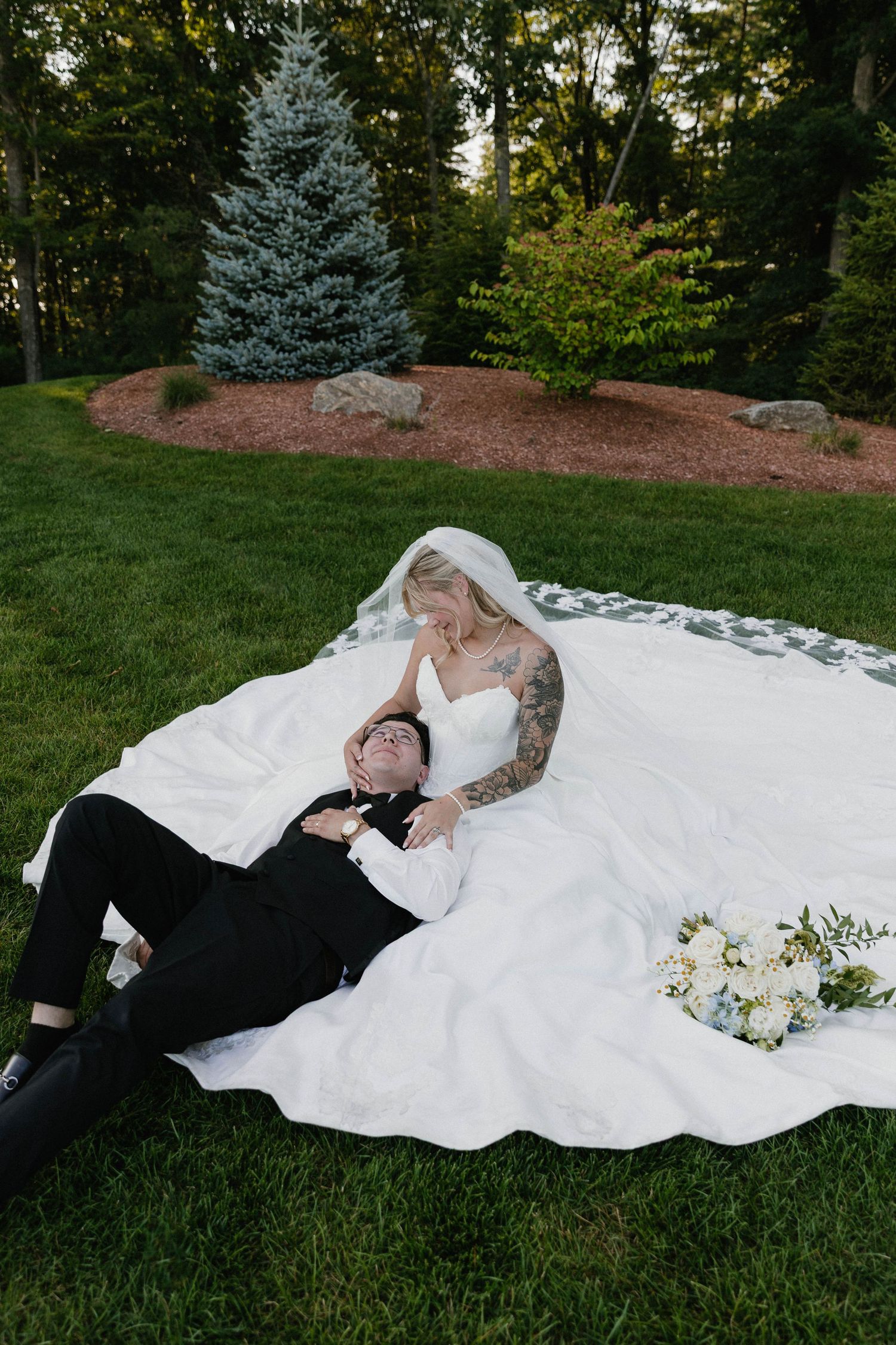 Bride and groom lying on white wedding dress on green grass, surrounded by trees, with a white floral bouquet nearby.