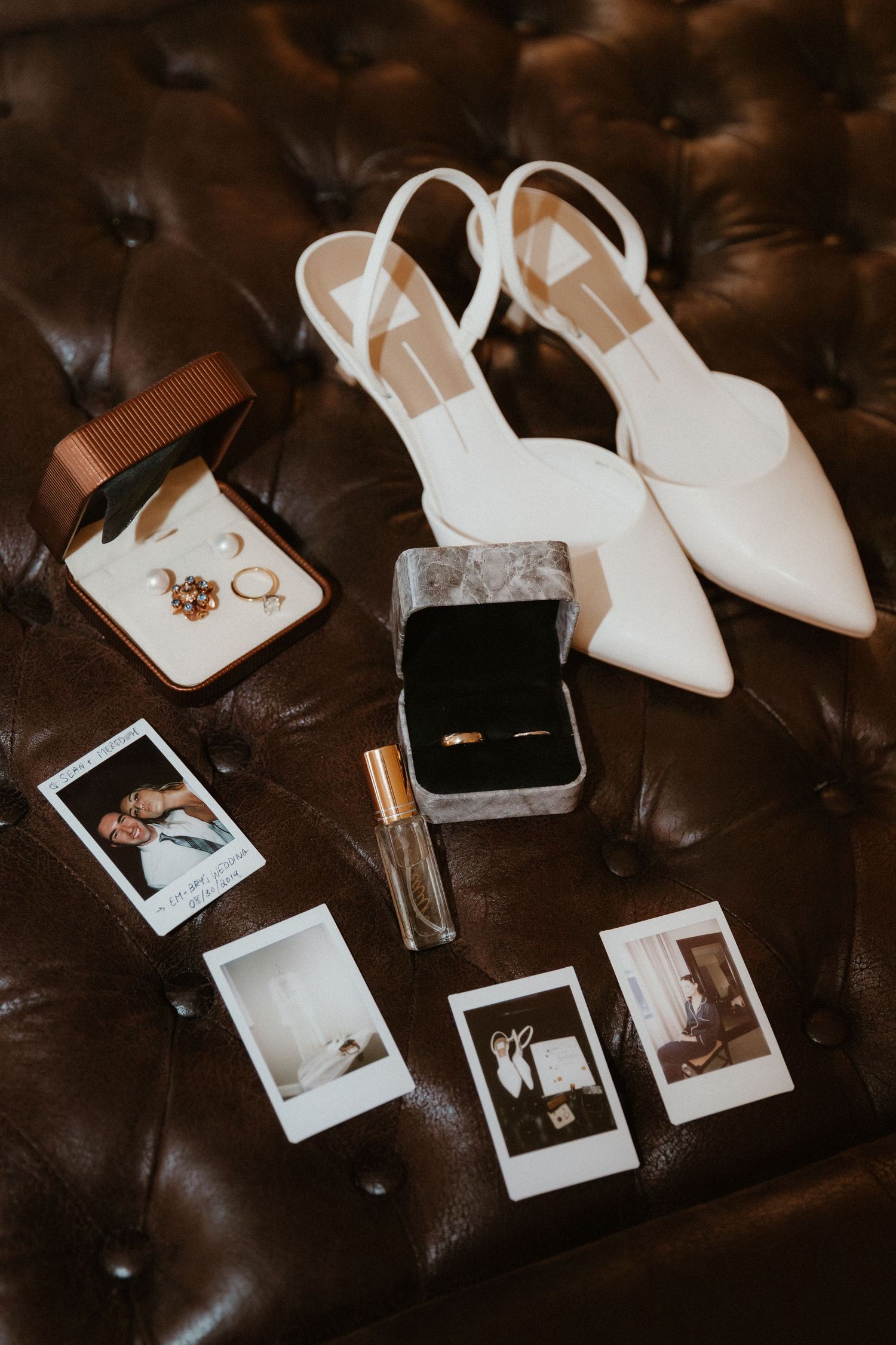 White bridal shoes and wedding details arranged on dark leather surface with scattered polaroid photographs.
