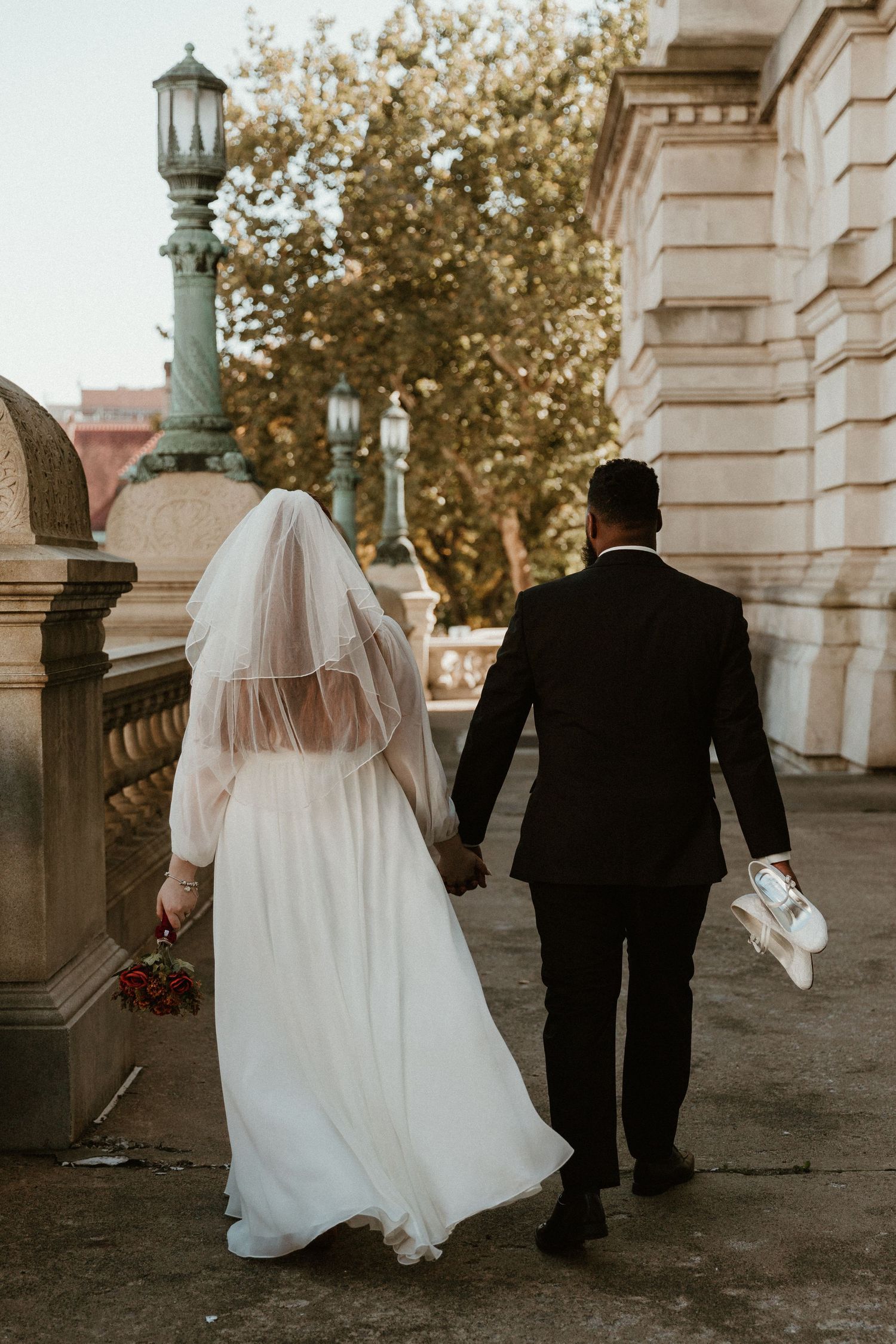 Newlyweds walk hand in hand along historic building with ornate lamp post.