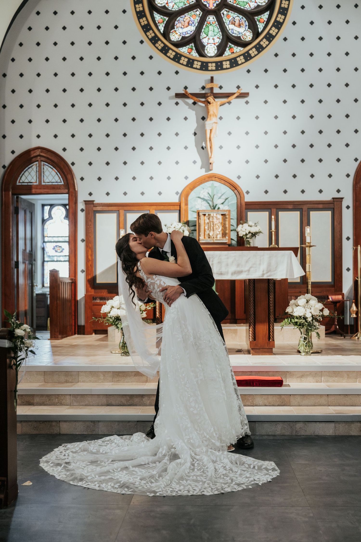 Wedding couple shares romantic moment in church with ornate stained glass window and decorative altar.