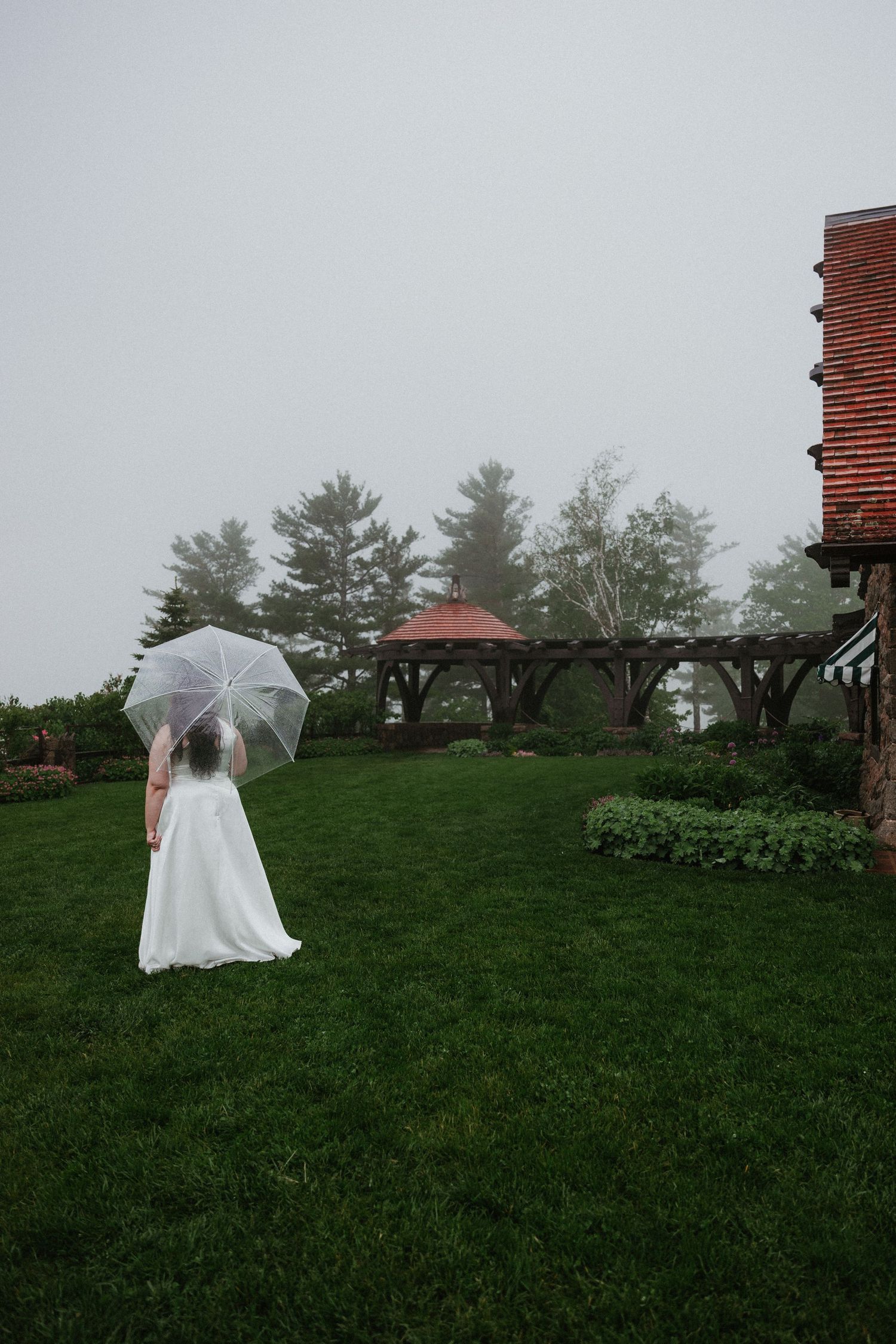 White umbrella and wedding dress against misty green landscape and stone building.