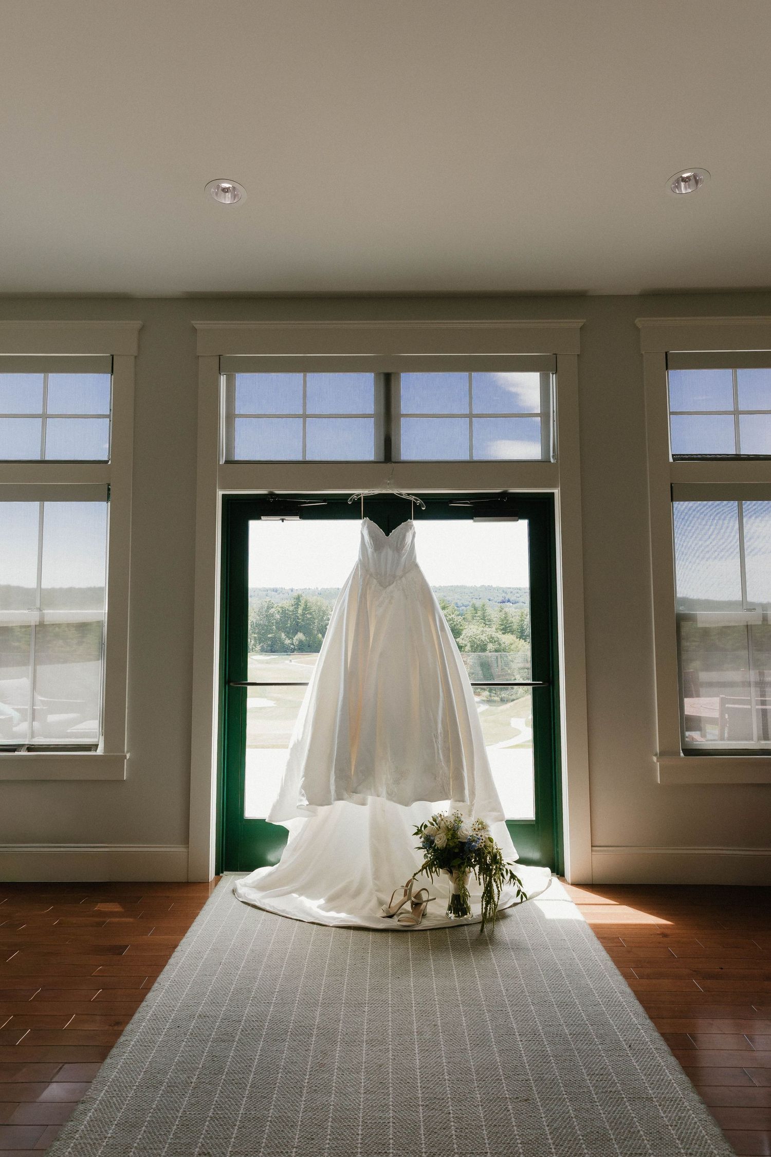 White wedding dress hanging in sunlit window with floral bouquet on floor below, elegant bridal gown display.