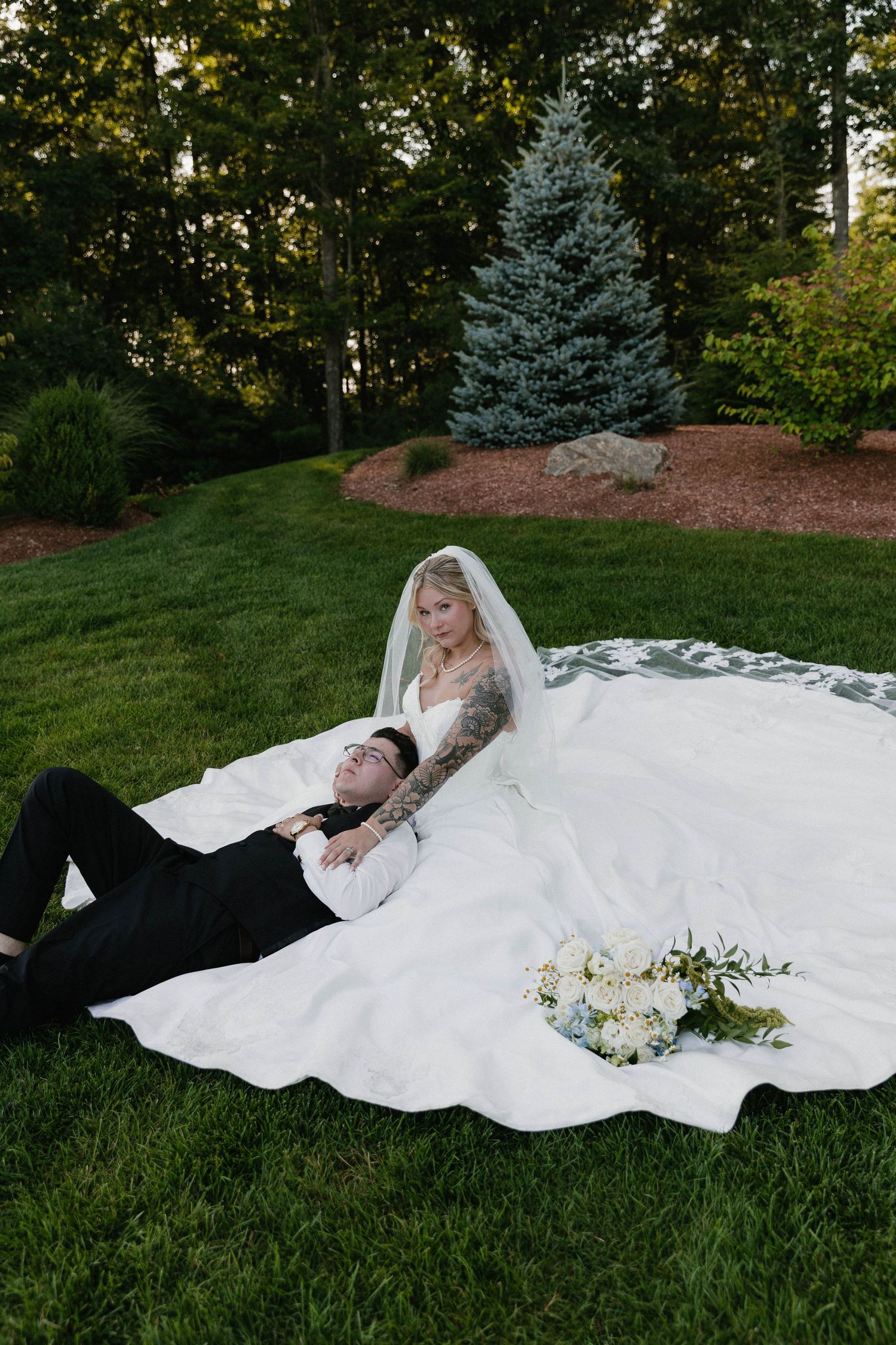 Bride in white gown and veil sits on grass while groom in black suit rests his head on her lap outdoors.
