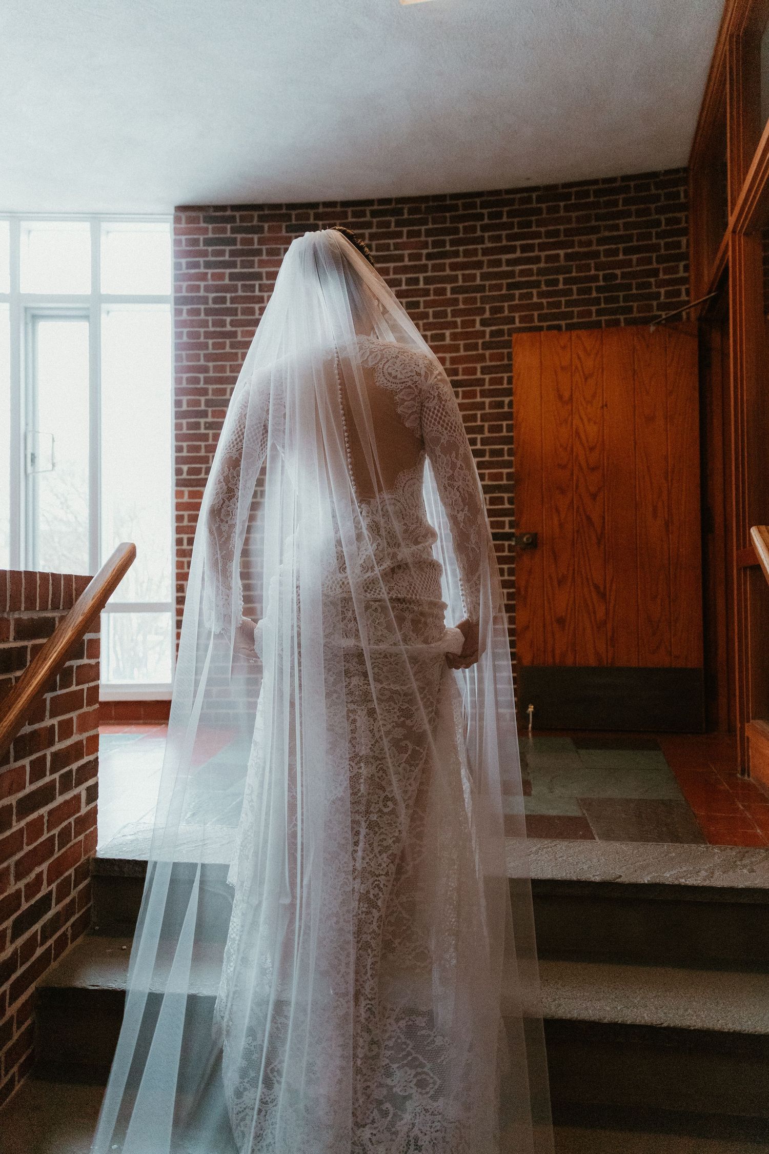 A bride in a lace wedding dress and long veil stands on a staircase against exposed brick walls.