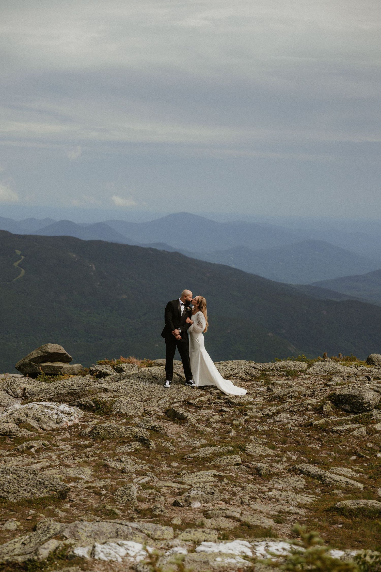 A couple shares an intimate moment on a rocky mountain summit with sweeping mountain views.