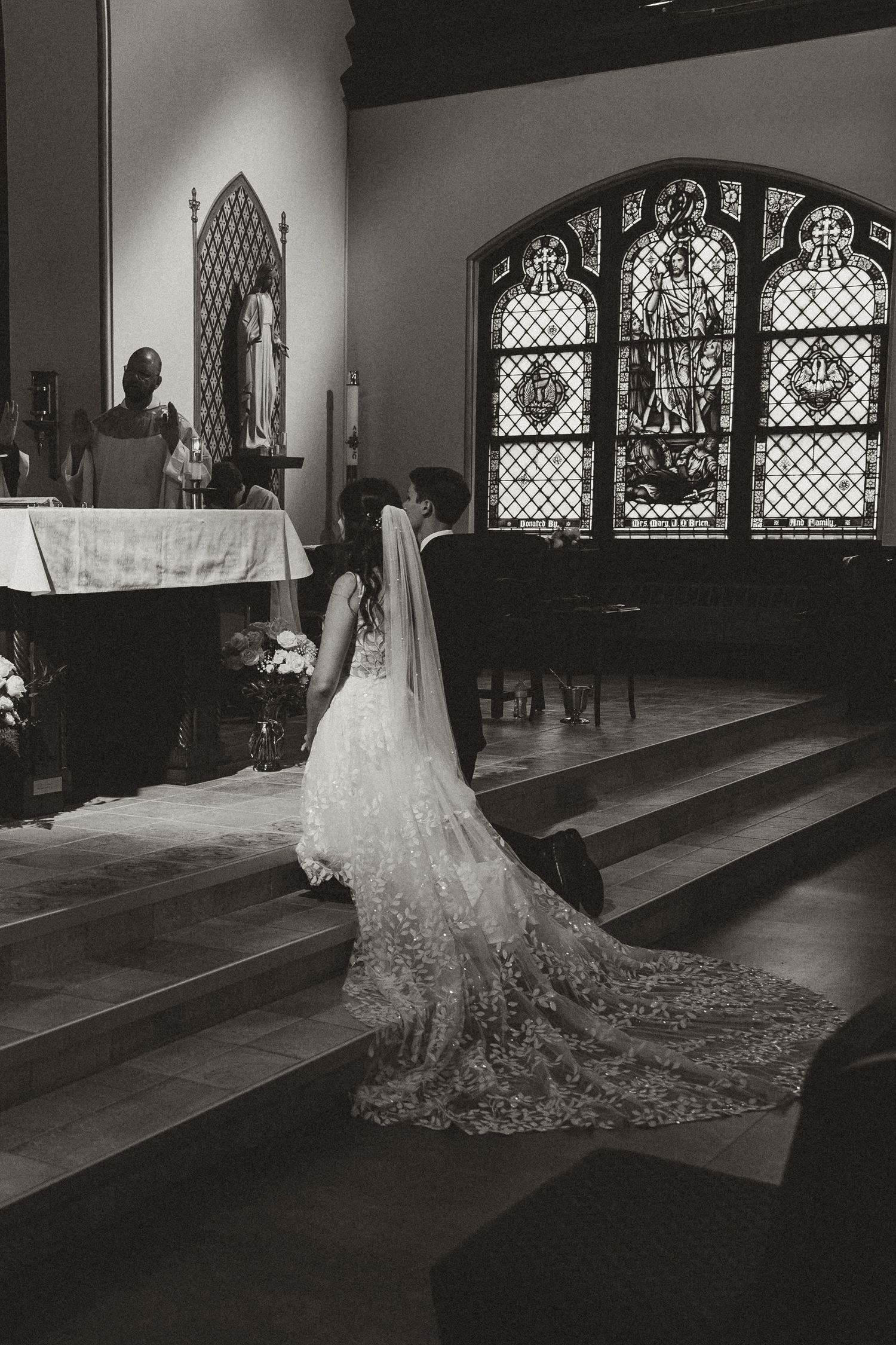 A dramatic black and white wedding photo showing a bride in a long lace train gown inside a church with stained glass windows.