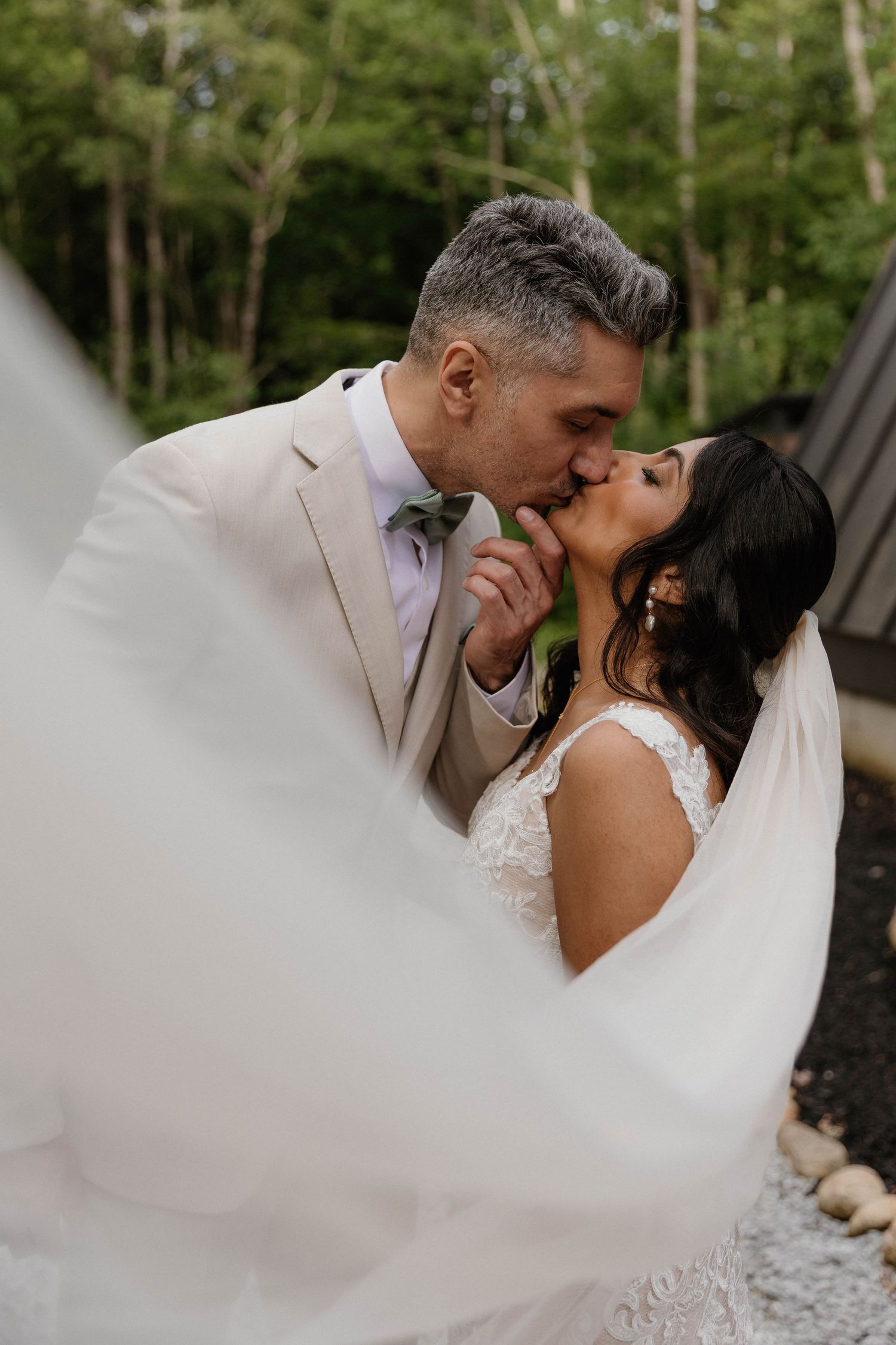 A bride and groom share a romantic kiss outdoors, her veil flowing in the foreground, surrounded by lush green trees.