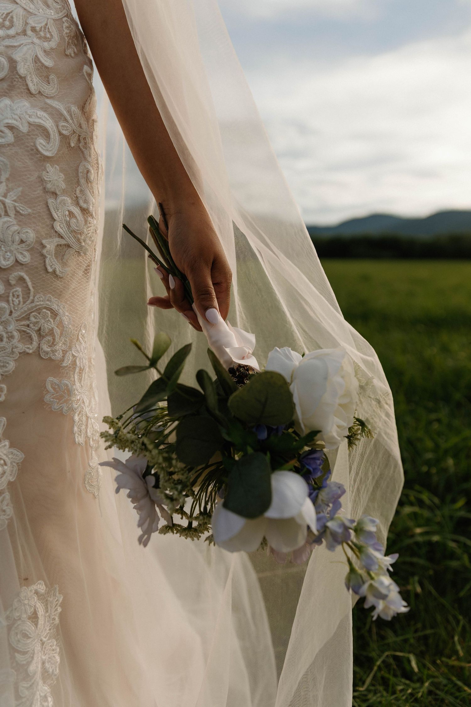 Bride holding a white and blue floral bouquet with greenery, wearing a lace wedding gown and veil in a field.