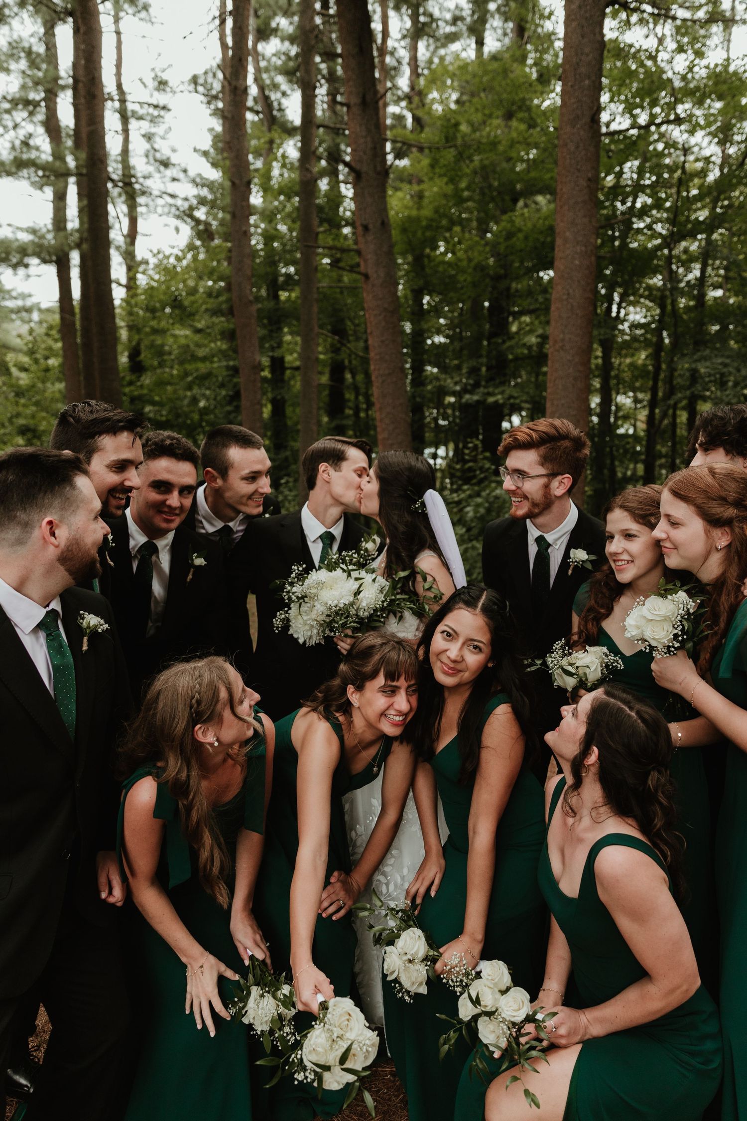 Wedding party in dark green dresses and black suits laughs together in a candid moment among tall trees.