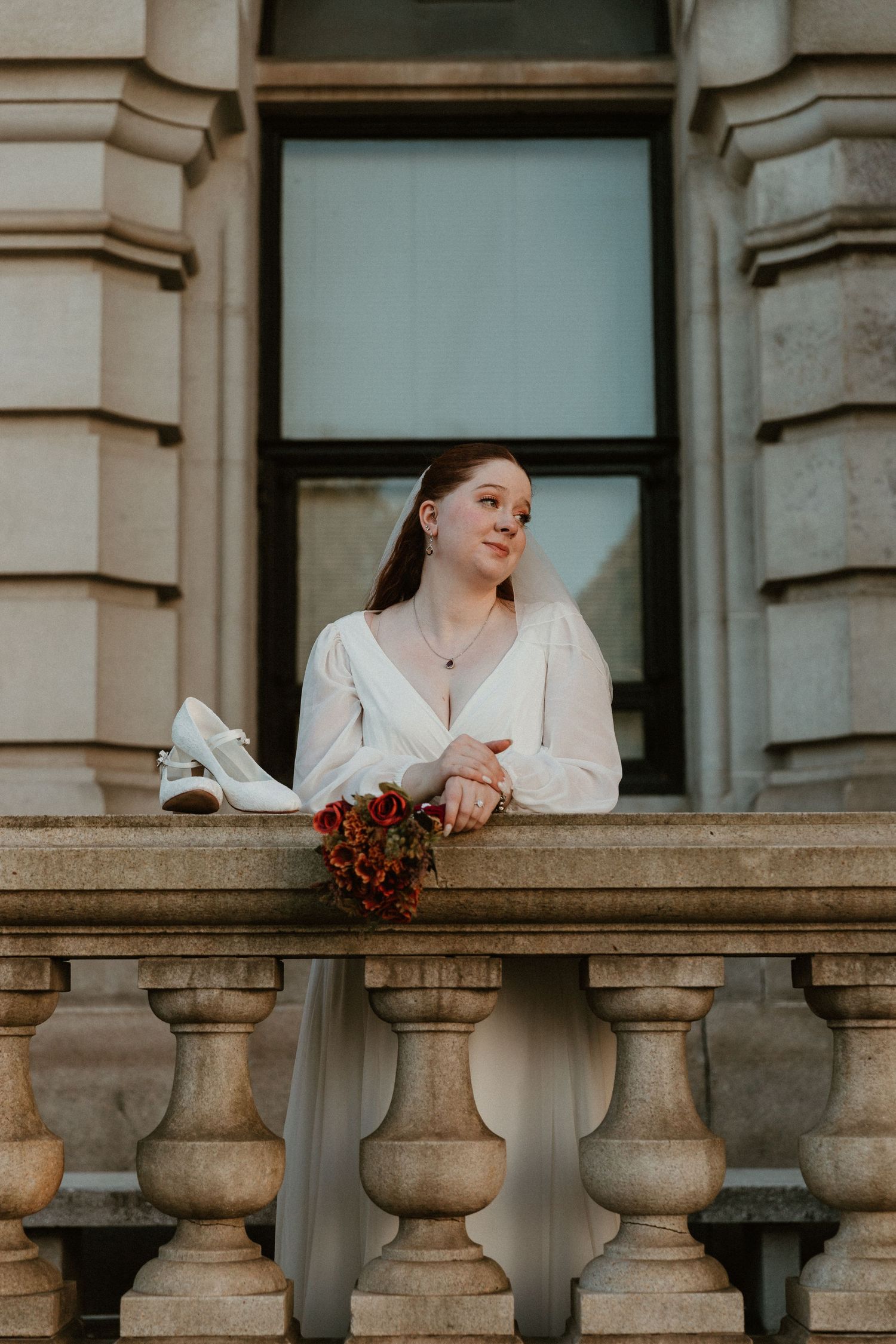 A bride in white holds red flowers while standing at an ornate stone balcony railing.