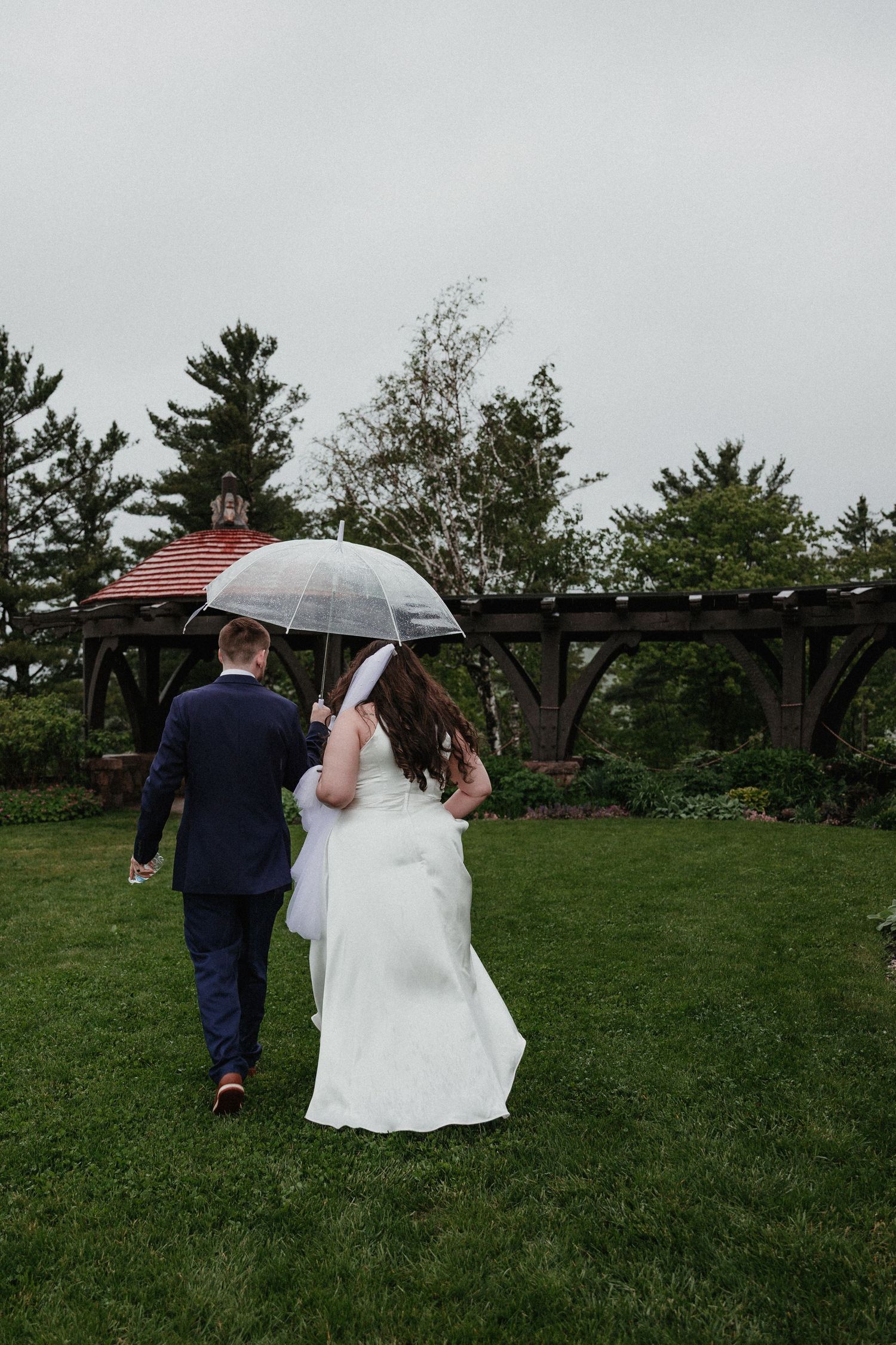 Wedding couple walking under clear umbrella on rainy day across green lawn.