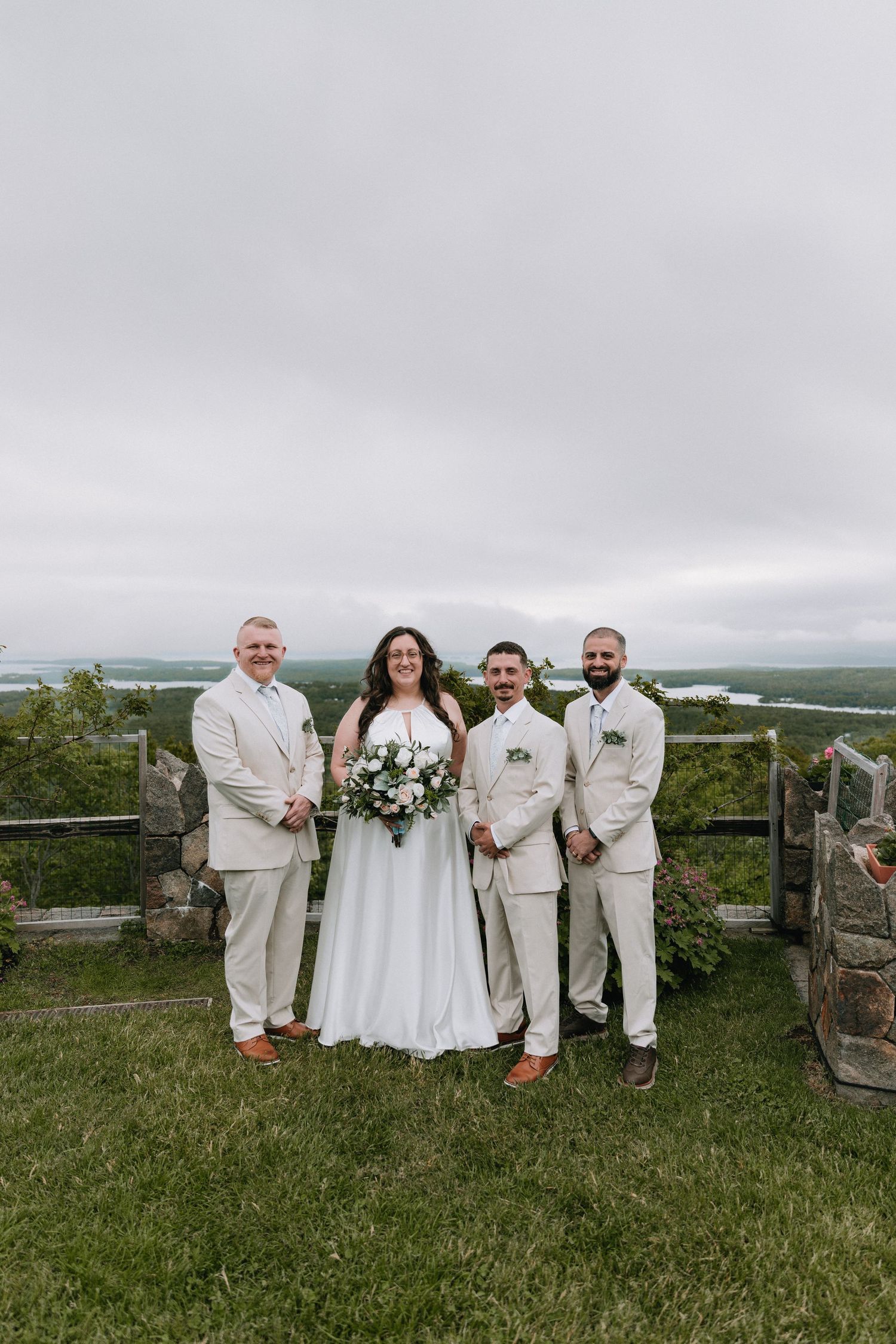 Wedding party in white and cream attire poses on grassy hilltop with scenic mountain backdrop.