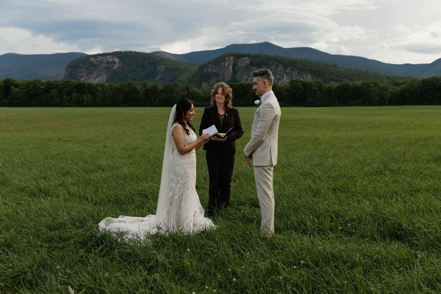 Outdoor wedding ceremony in a green field with mountain backdrop, bride in white gown, groom in tan suit, officiant holding book.