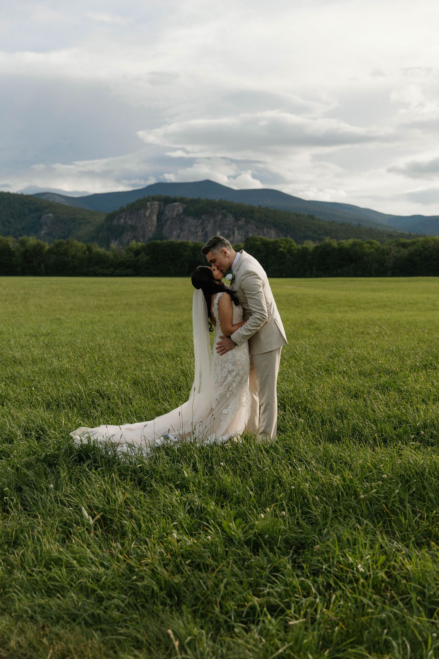 Bride and groom embracing in a green field with mountain backdrop on their wedding day.