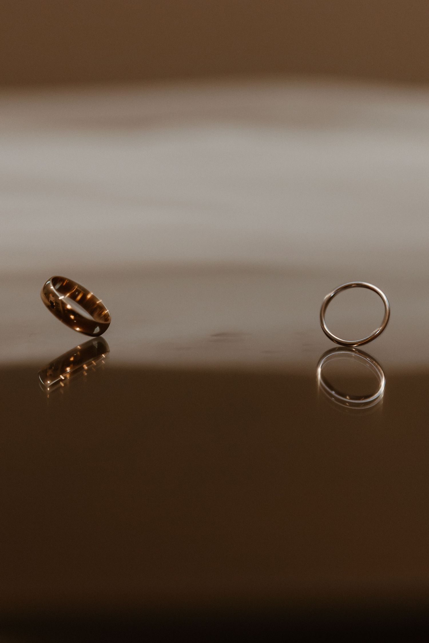 Two wedding rings reflecting on a glossy dark surface in moody lighting.