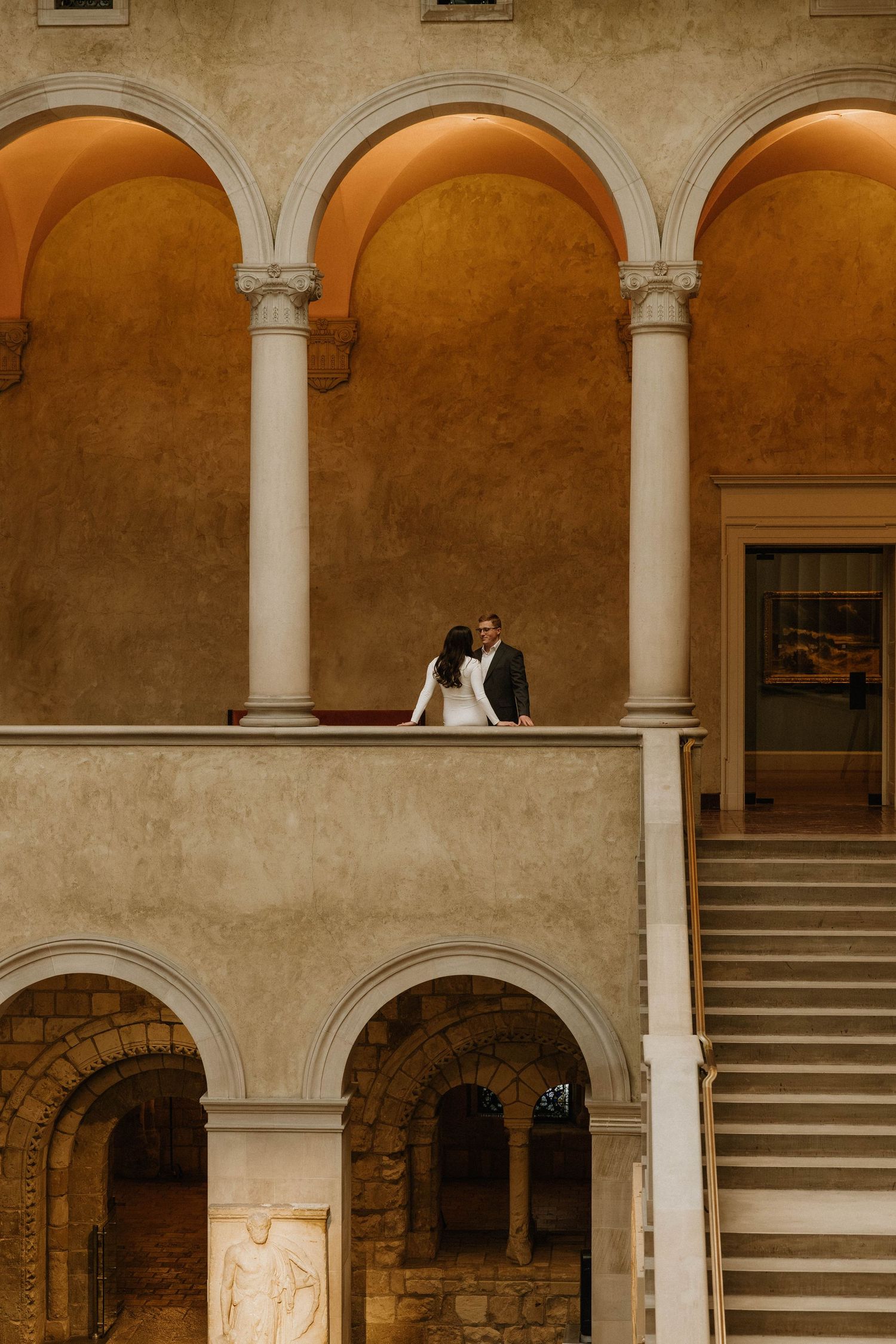 A couple stands in an elegant arched courtyard with stone columns and stairs creating a romantic architectural setting.
