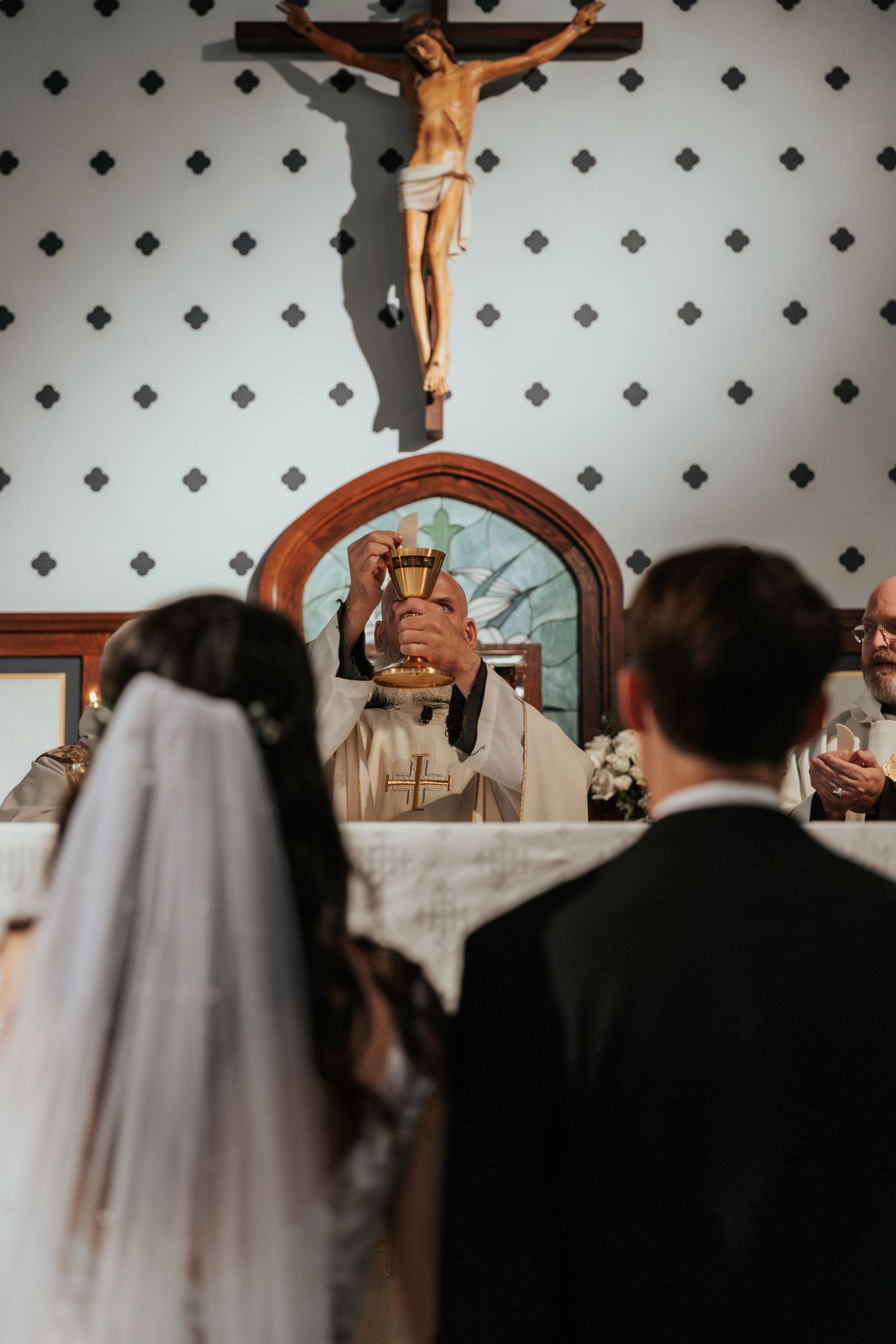 A Catholic wedding ceremony taking place in a church with a crucifix mounted on a patterned wall.