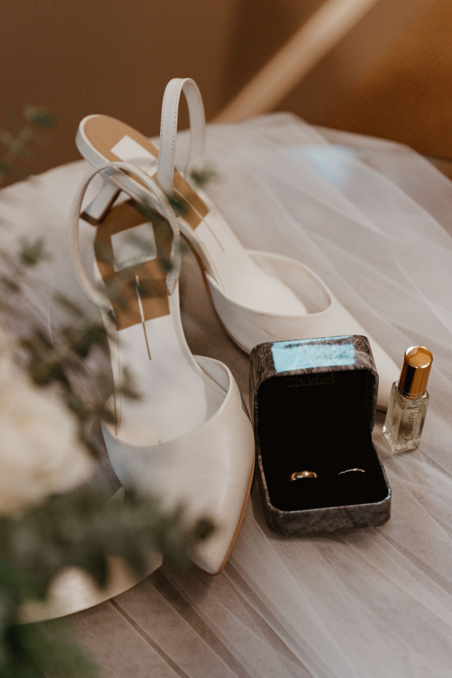 White bridal shoes, ring box, and perfume bottle arranged on wooden surface with veil.