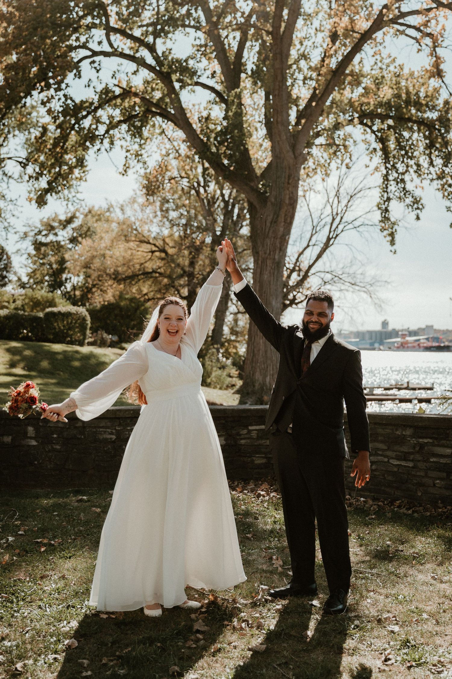 A joyful wedding couple holds hands raised under a large tree in an outdoor autumn setting.