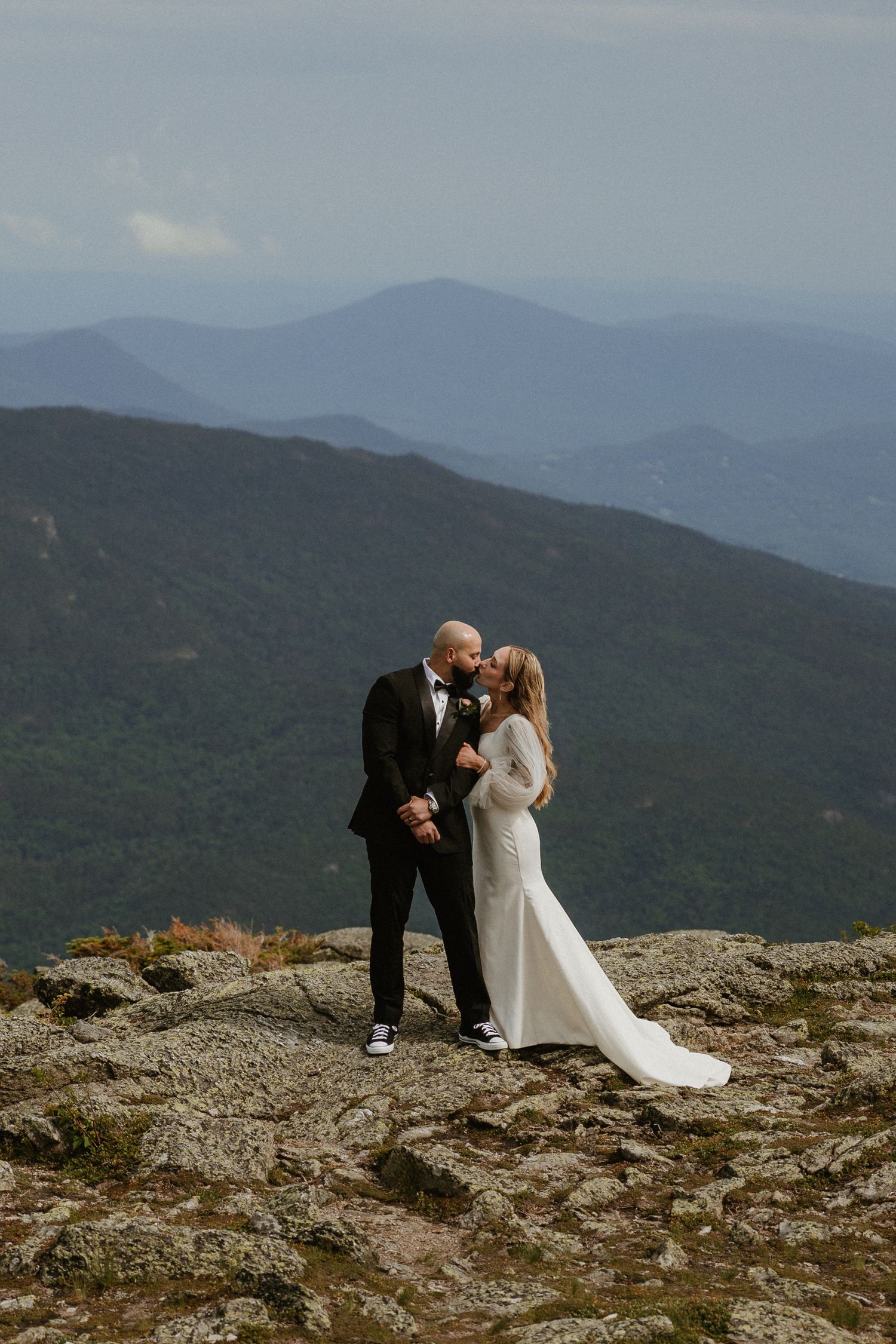 Newlyweds embrace on a rocky mountain summit with sweeping mountain ranges in the background.