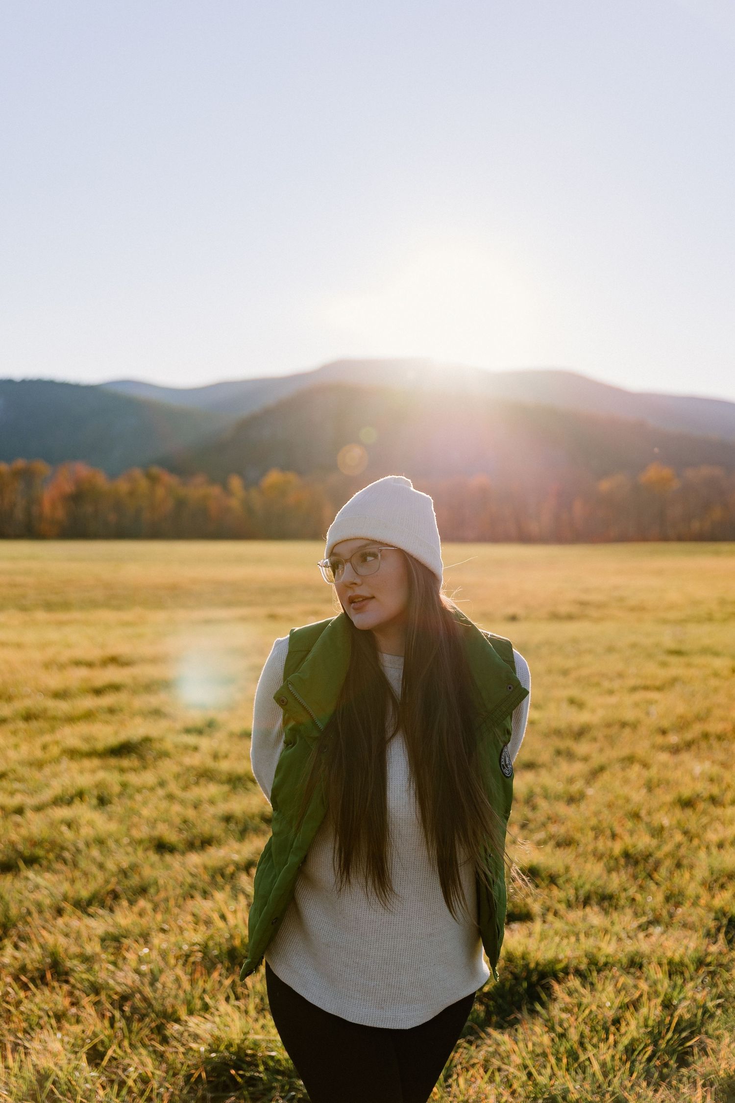 Person in a white beanie and green vest poses in golden field at sunset with mountains in background.