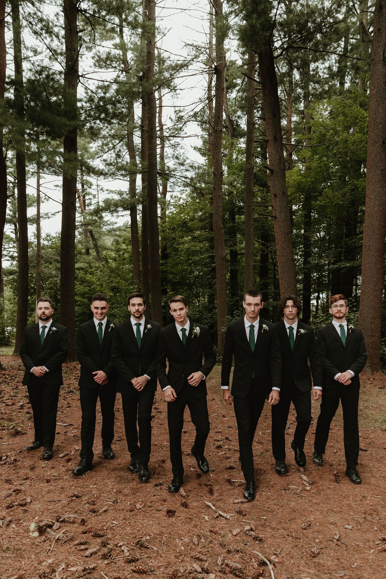 Groomsmen in black suits stand in formation on pine needle covered forest ground.