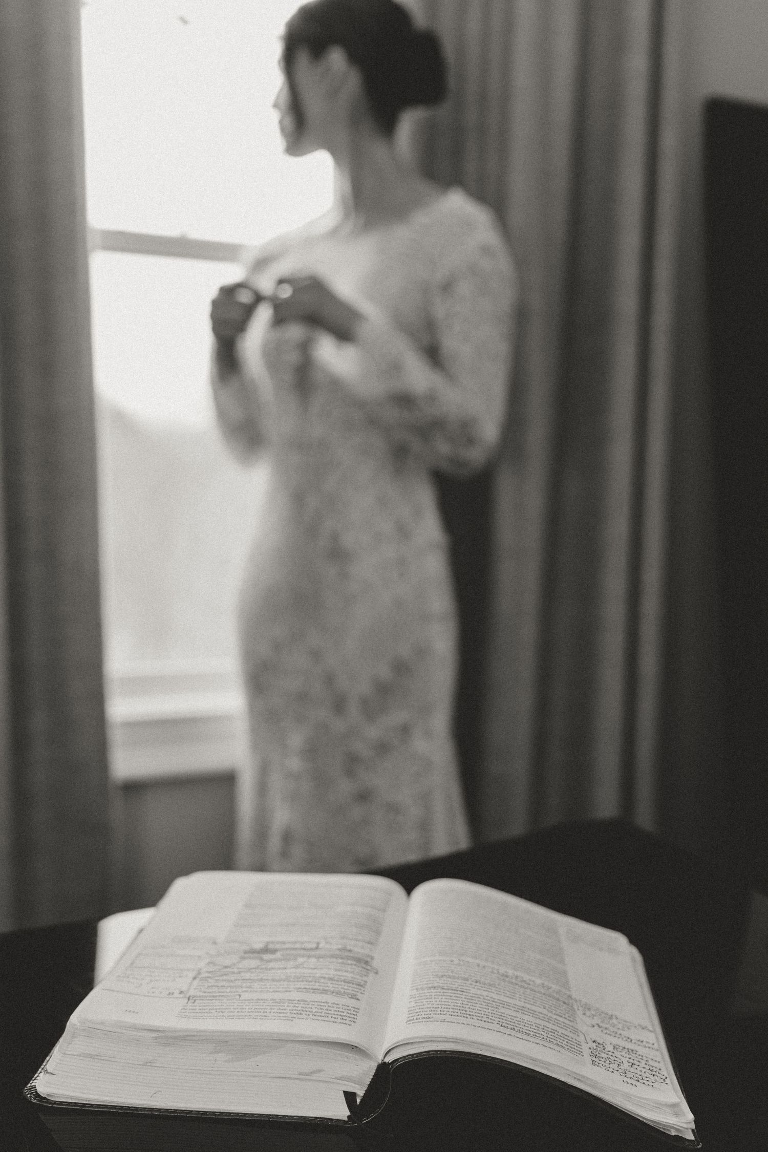 An open book sits on a table near a window with curtains while a person stands reading in soft natural light.