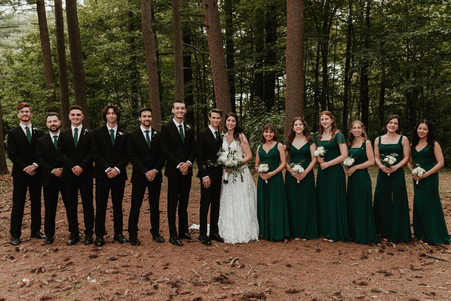 Wedding party in formal attire poses outdoors in a wooded setting with bridesmaids in emerald green dresses.